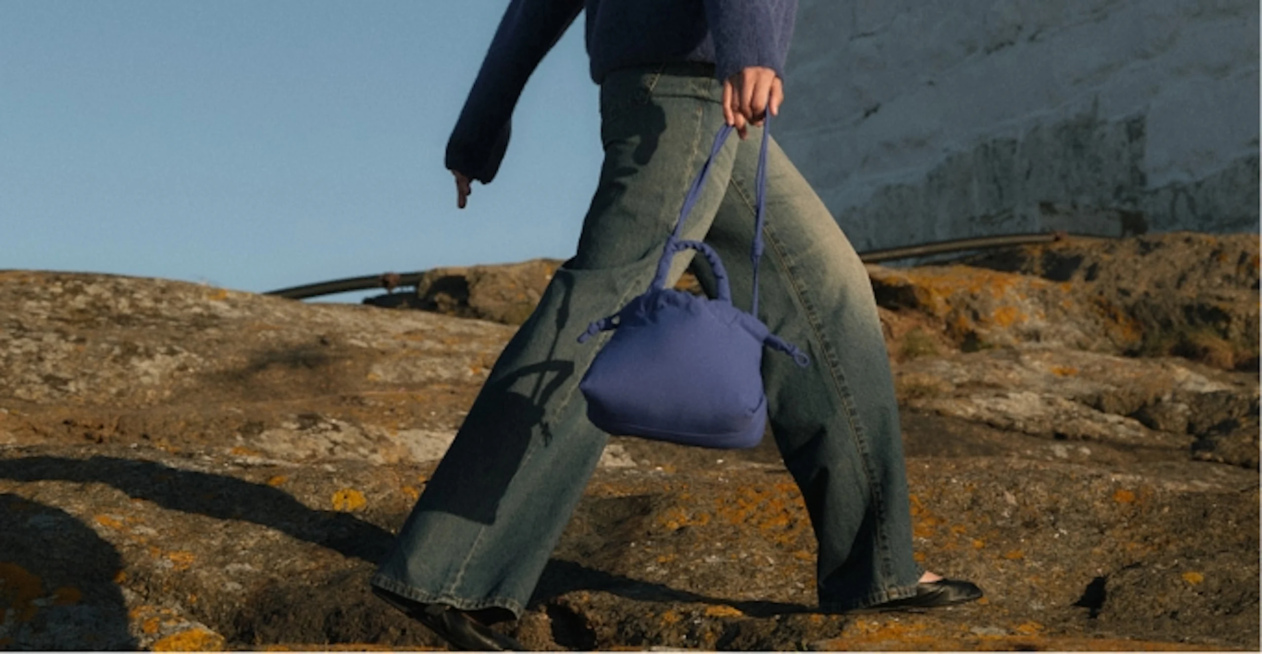 Person i blå genser og grå jeans går over klipper og bærer en liten blå pute-veske med flettet håndtak.