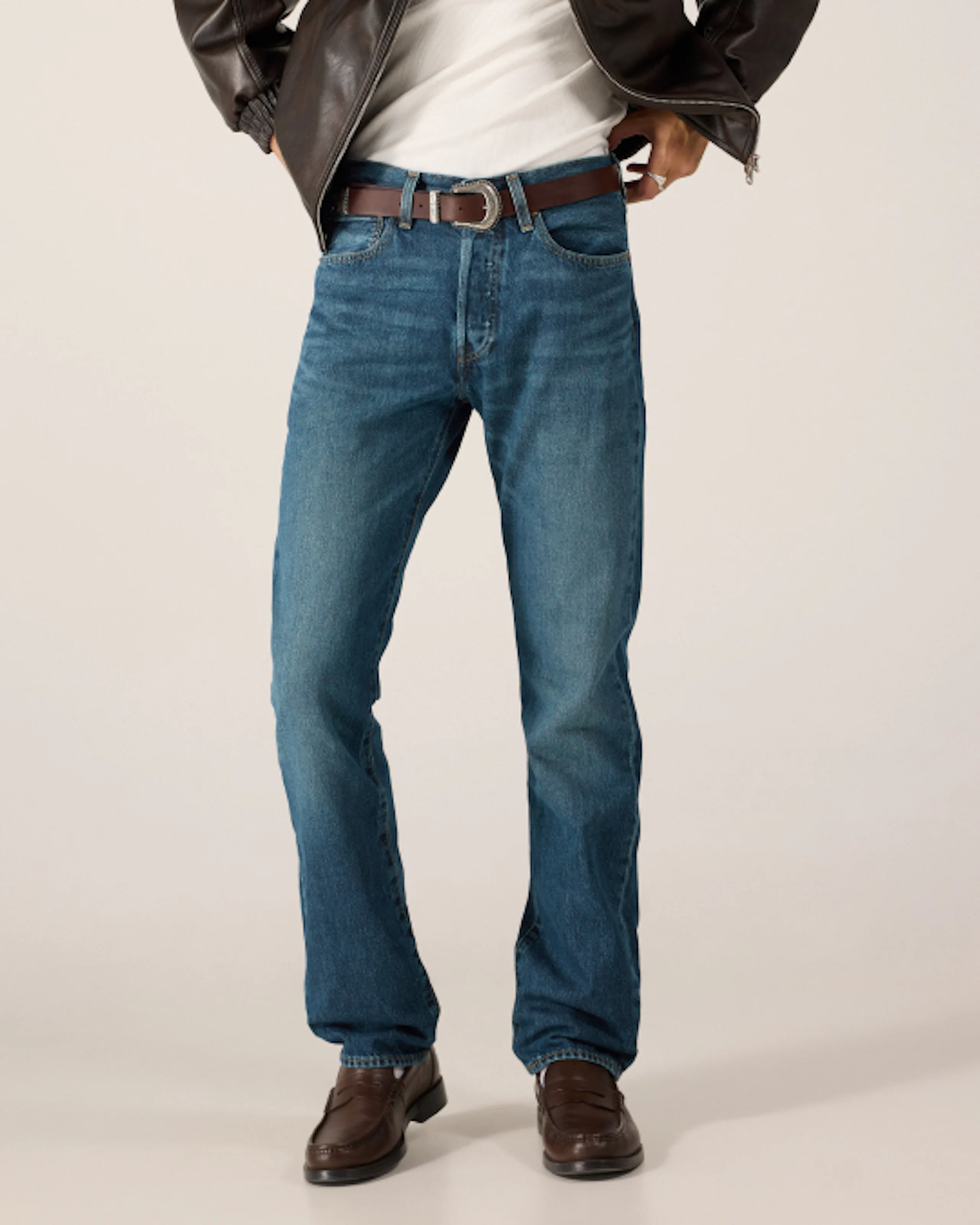 Man i mellanblå jeans, brunt läderbälte, vit t-shirt, mörkbrun jacka och bruna loafers.
