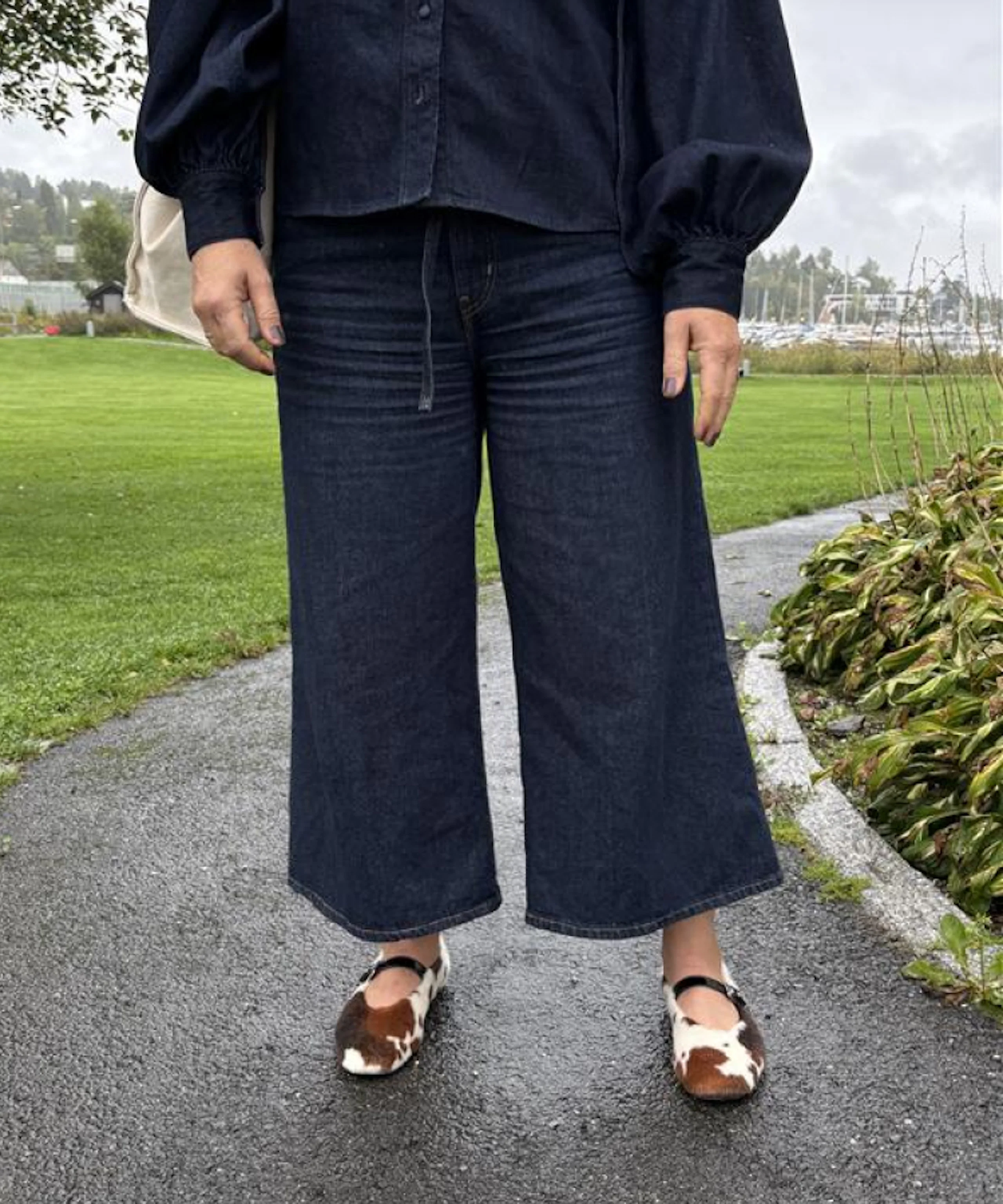 Person i mörkblå vida jeans och svart skjorta, med ko-mönstrade platta skor, står utomhus på en gångväg.