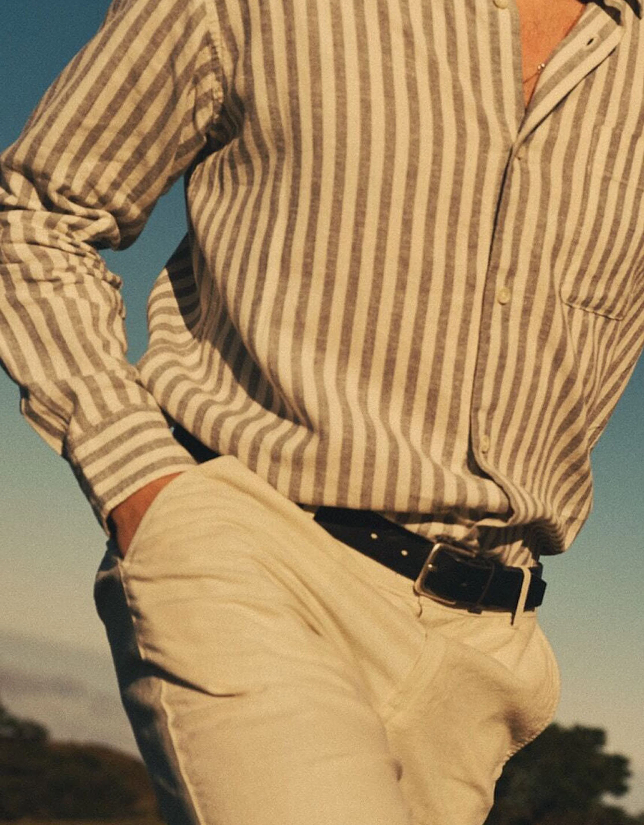 Person iført en stripete lin-skjorte i beige og grå, beige bukser og et svart belte, med hendene bak ryggen.