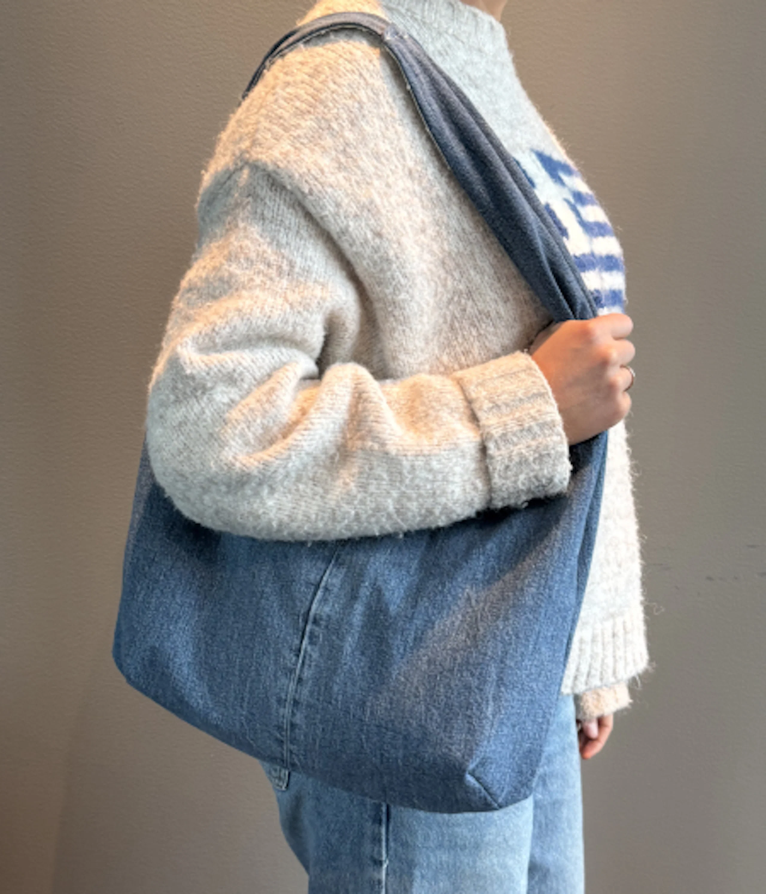 Person som har på seg en kremfarget genser og jeans og bærer en stor blå denim skulderveske over høyre skulder foran en nøytral vegg.