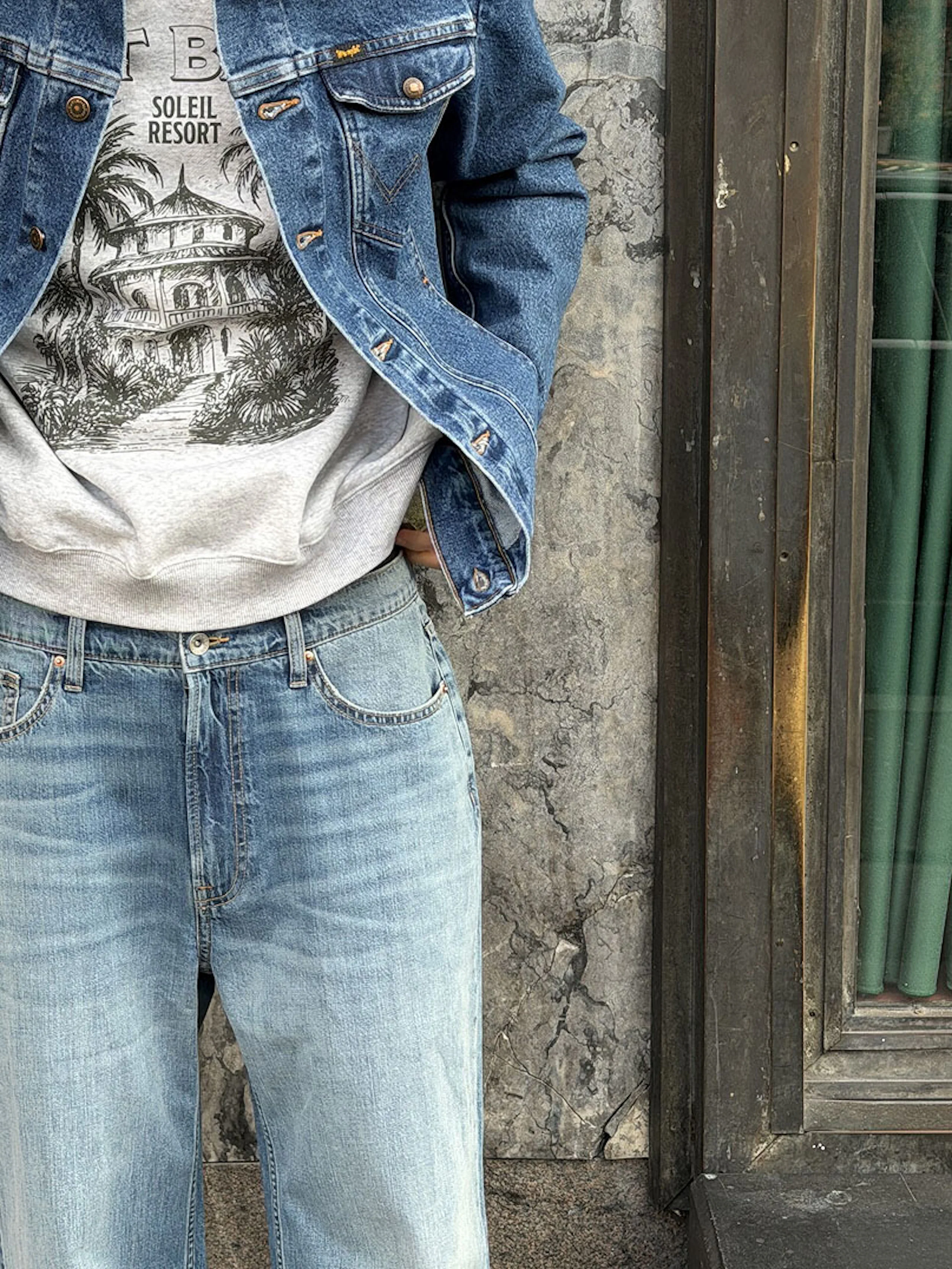 Person i blå jeansjacka, grå tröja med "Soleil Resort"-tryck och ljusblå jeans, står framför en stenvägg.