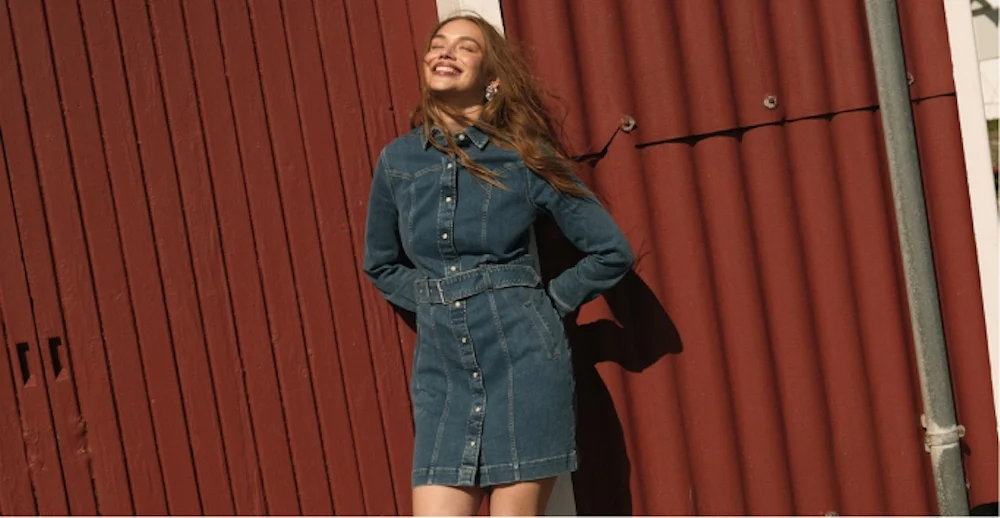 Smilende ung kvinne i blå denimkjole med belte, står foran rød vegg i sollys.