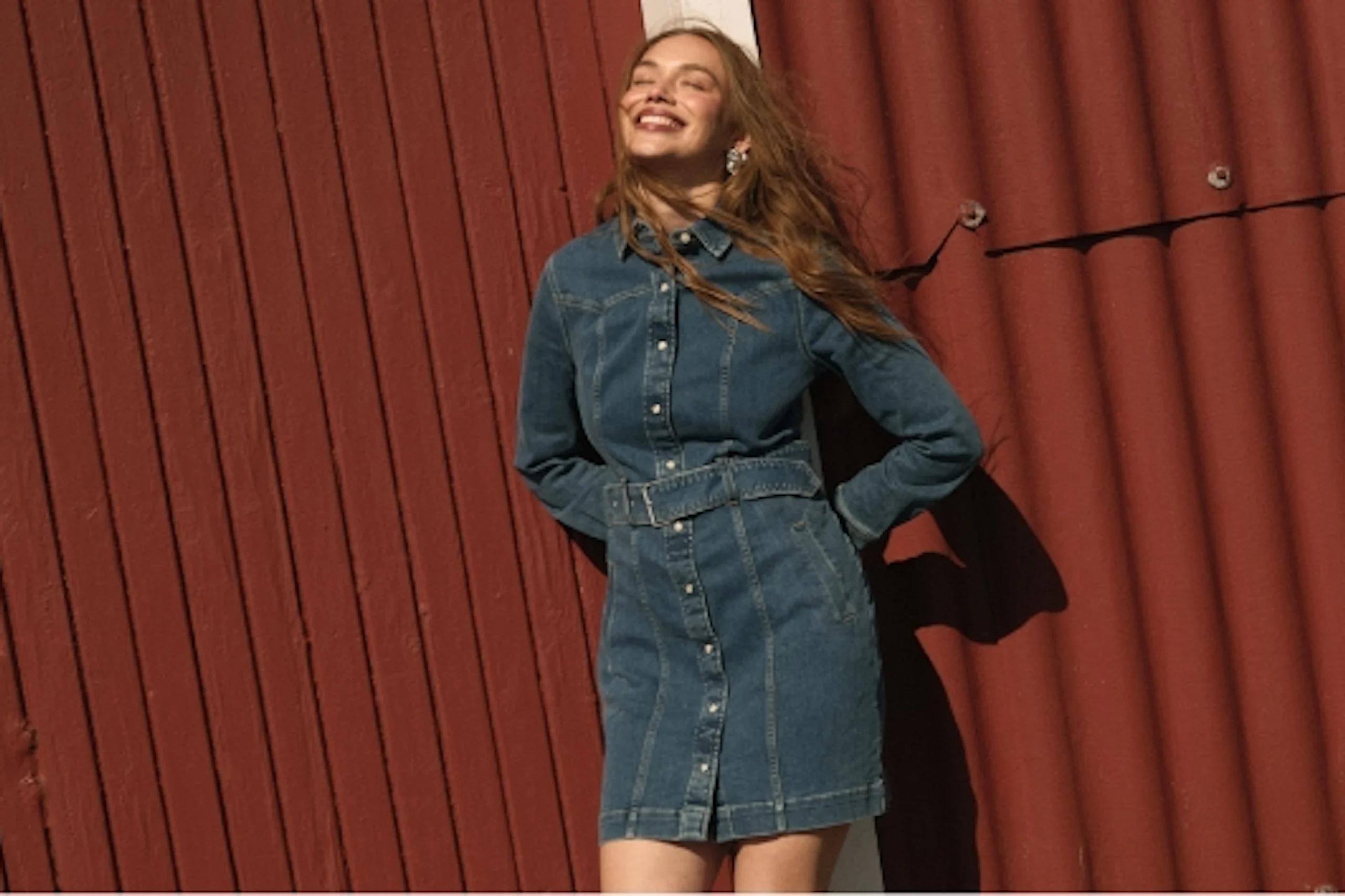 Smilende ung kvinne i blå denimkjole med belte, står foran rød vegg i sollys.