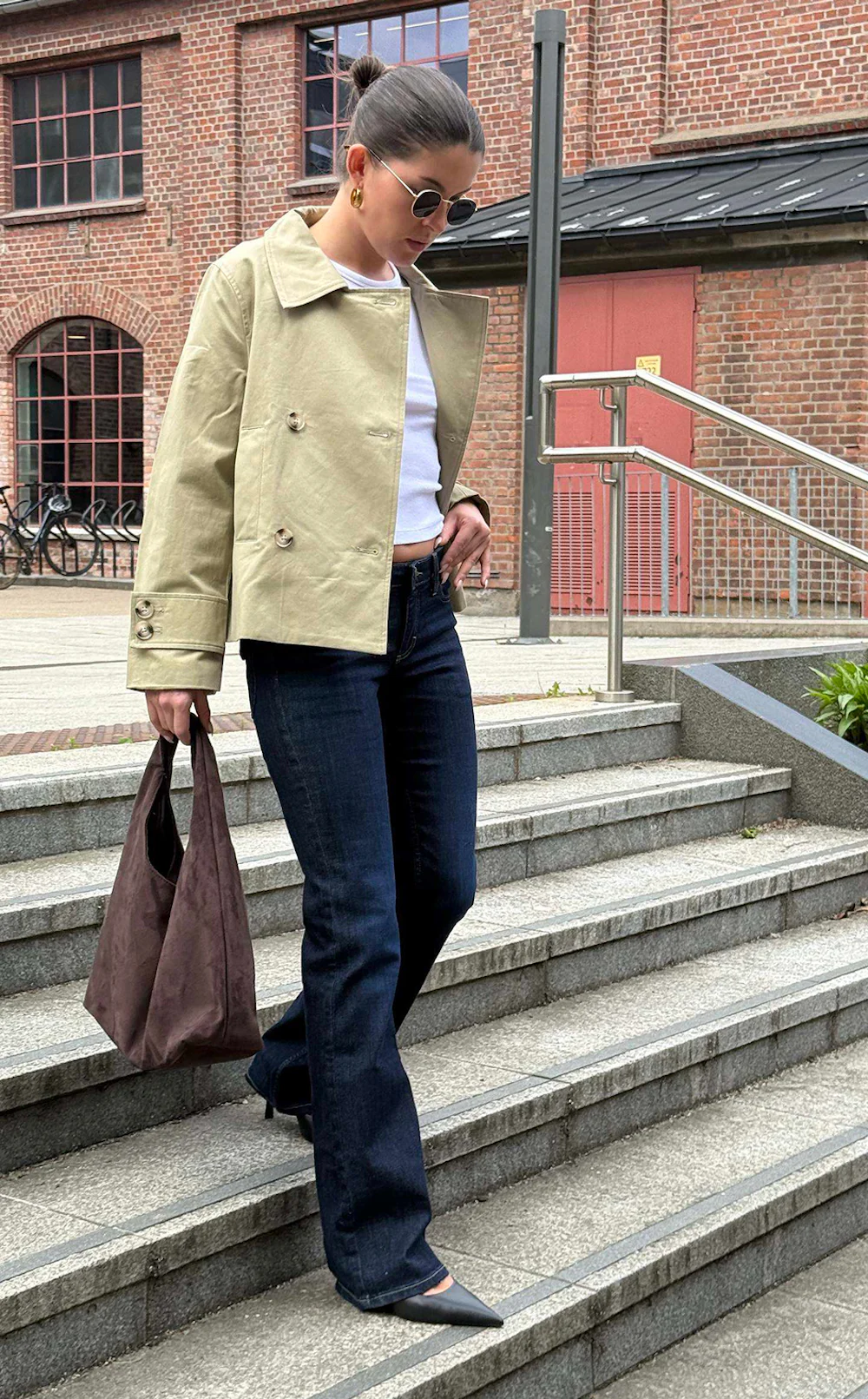 Kvinne som går ned utendørs trapp iført beige jakke, mørke utsvingte jeans, svarte høyhælte sko og bærer en brun veske.