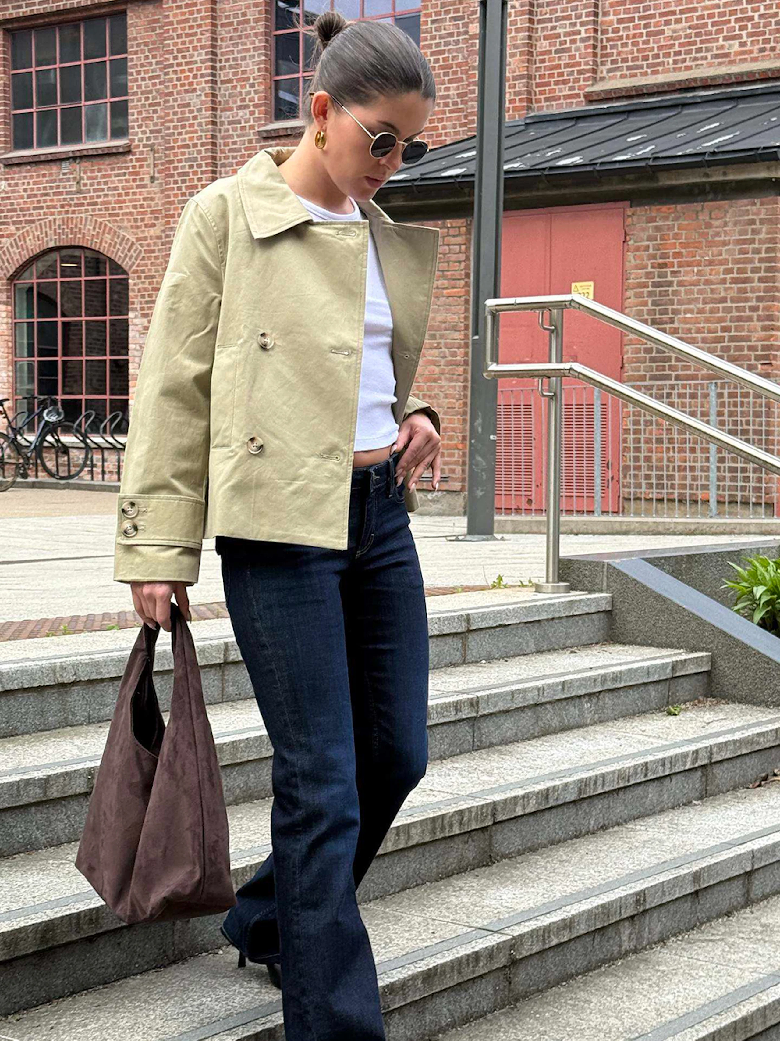 Kvinne som går ned utendørs trapp iført beige jakke, mørke utsvingte jeans, svarte høyhælte sko og bærer en brun veske.