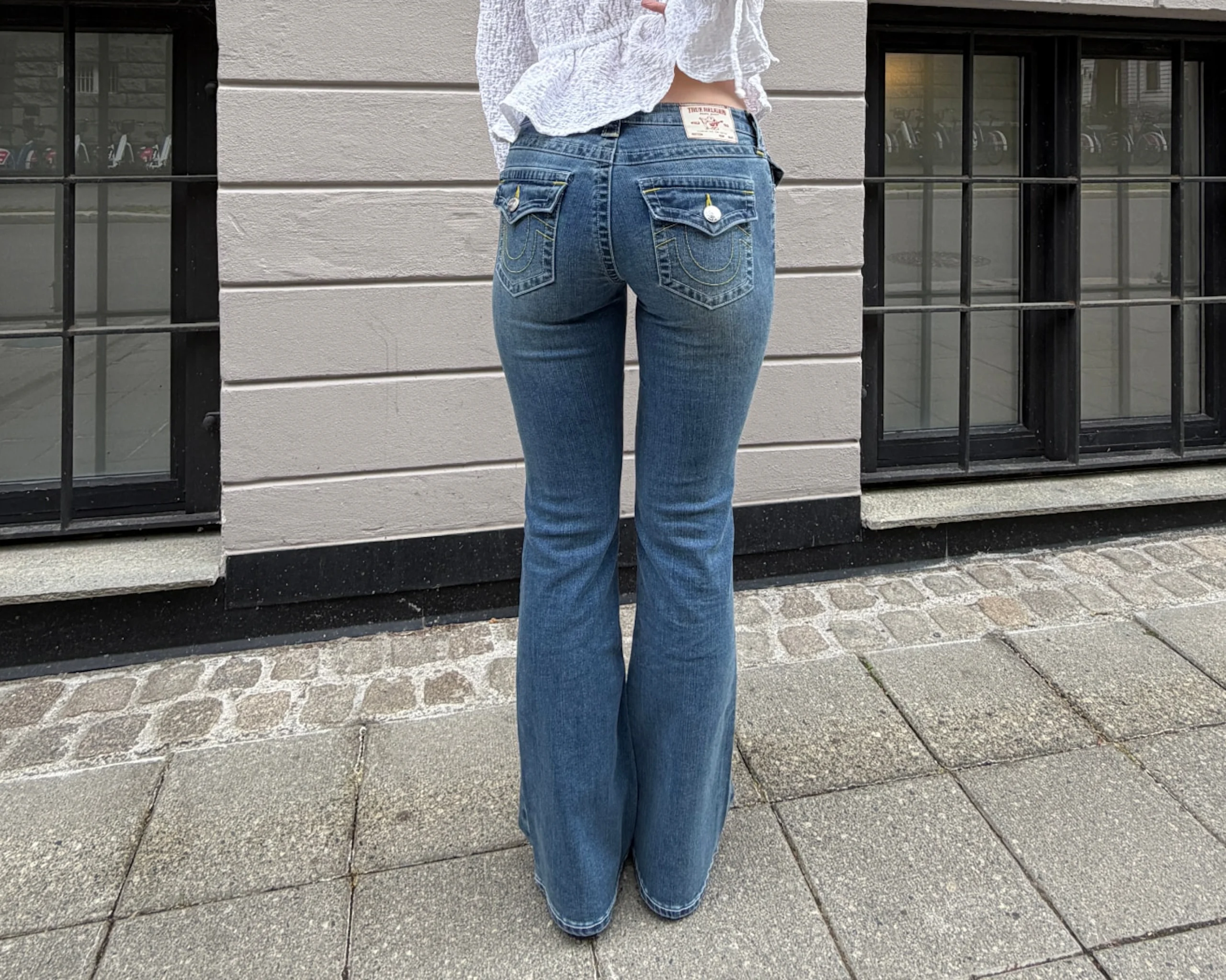 Bakifrån av en person som bär högmidjade utställda blå jeans med dekorativa bakfickor, stående på en kullerstensgata.