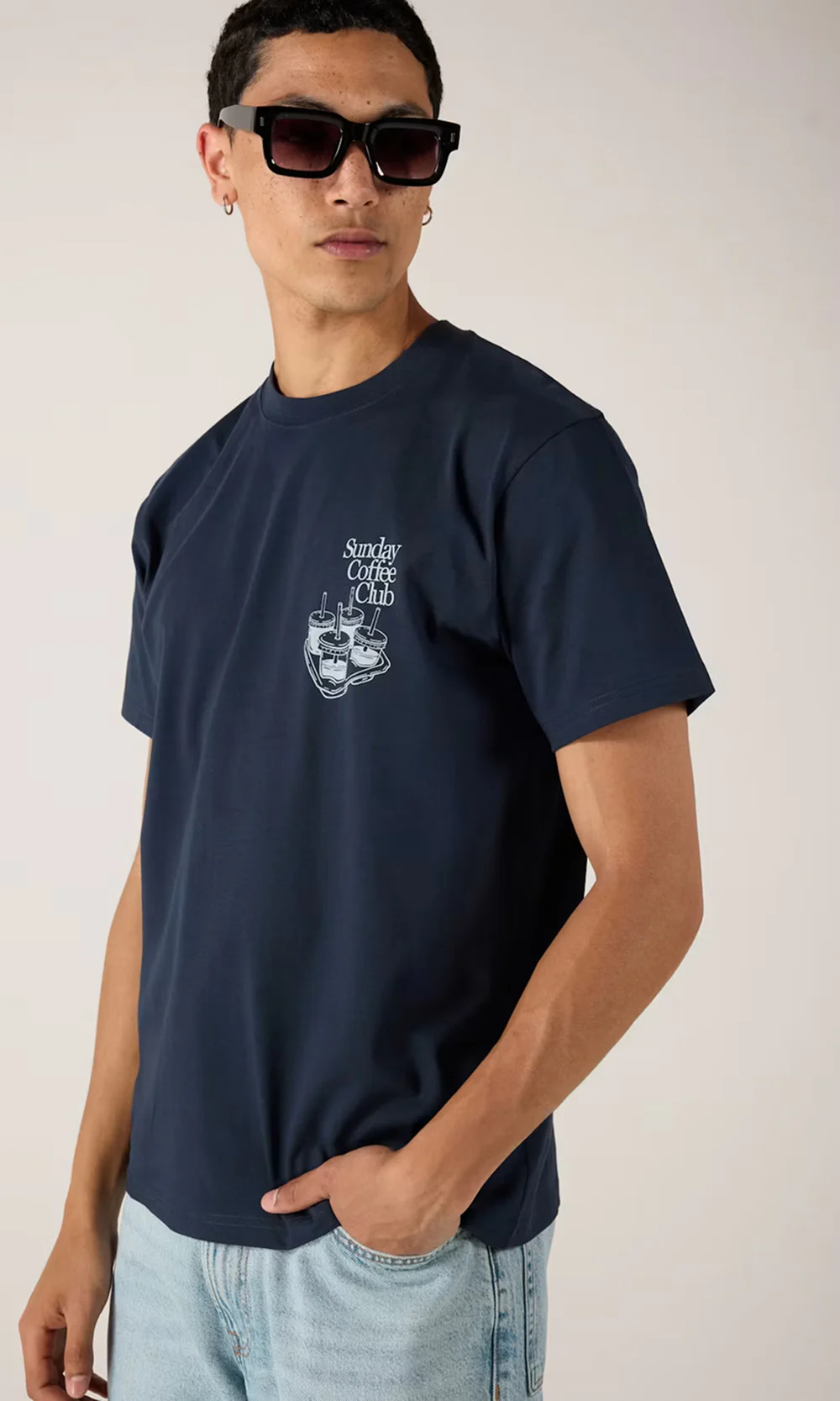 Man i marinblå t-shirt med "Sunday Coffee Club"-tryck, solglasögon och ljusblå jeans, med handen i fickan.