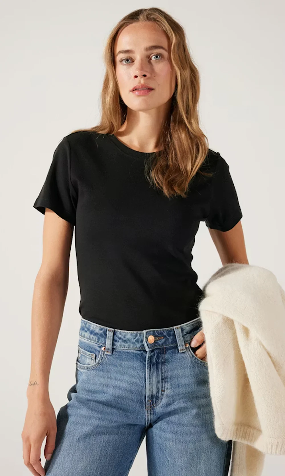 Ung kvinne iført en tettsittende svart T‑skjorte og blå jeans, holder en kremfarget strikkegenser over den ene armen, nøytral studiobakgrunn.