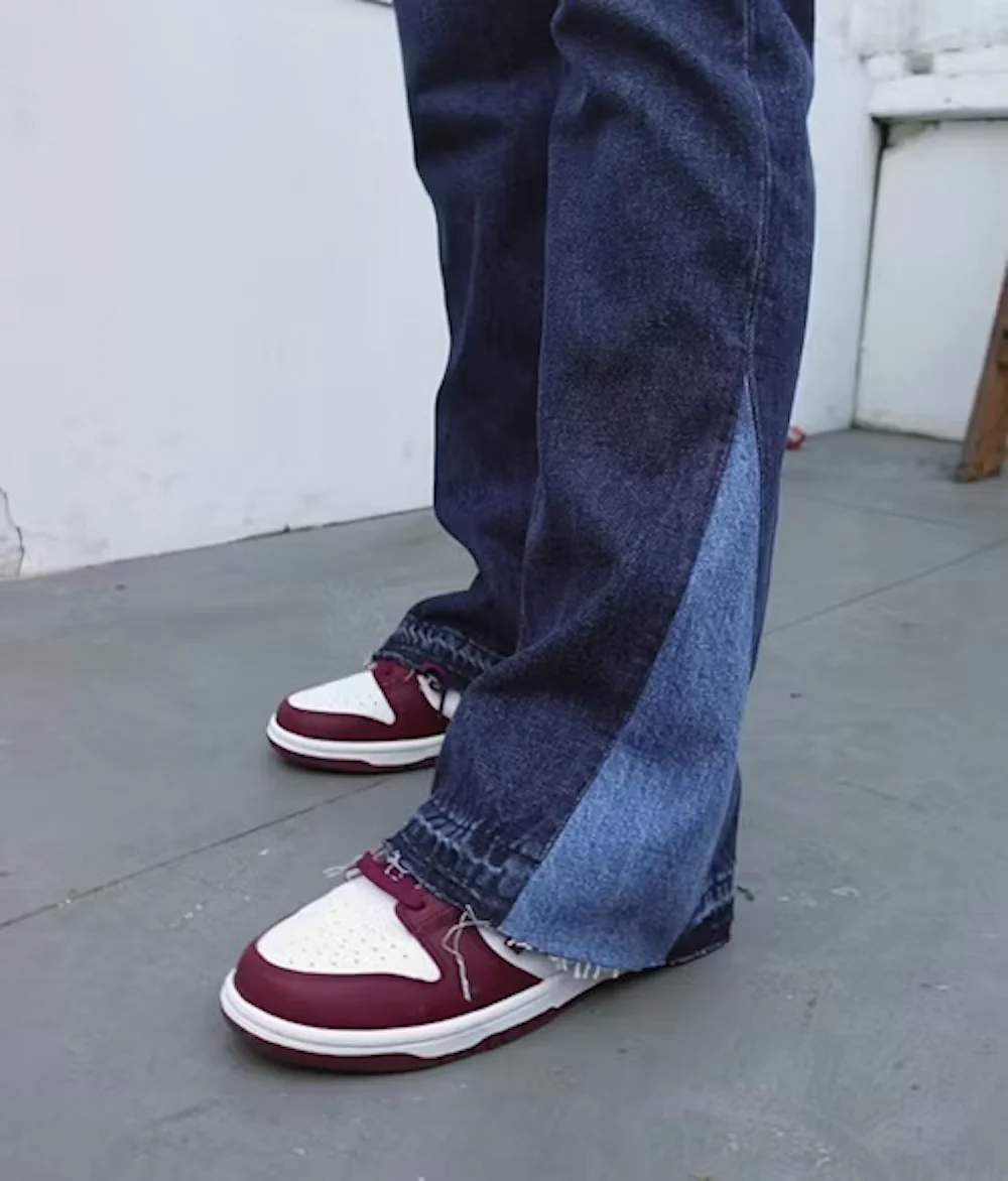 Lavvinklet bilde av tofargede blå jeans med rå kant som dekker burgunderrøde og hvite joggesko på et grått fortau.