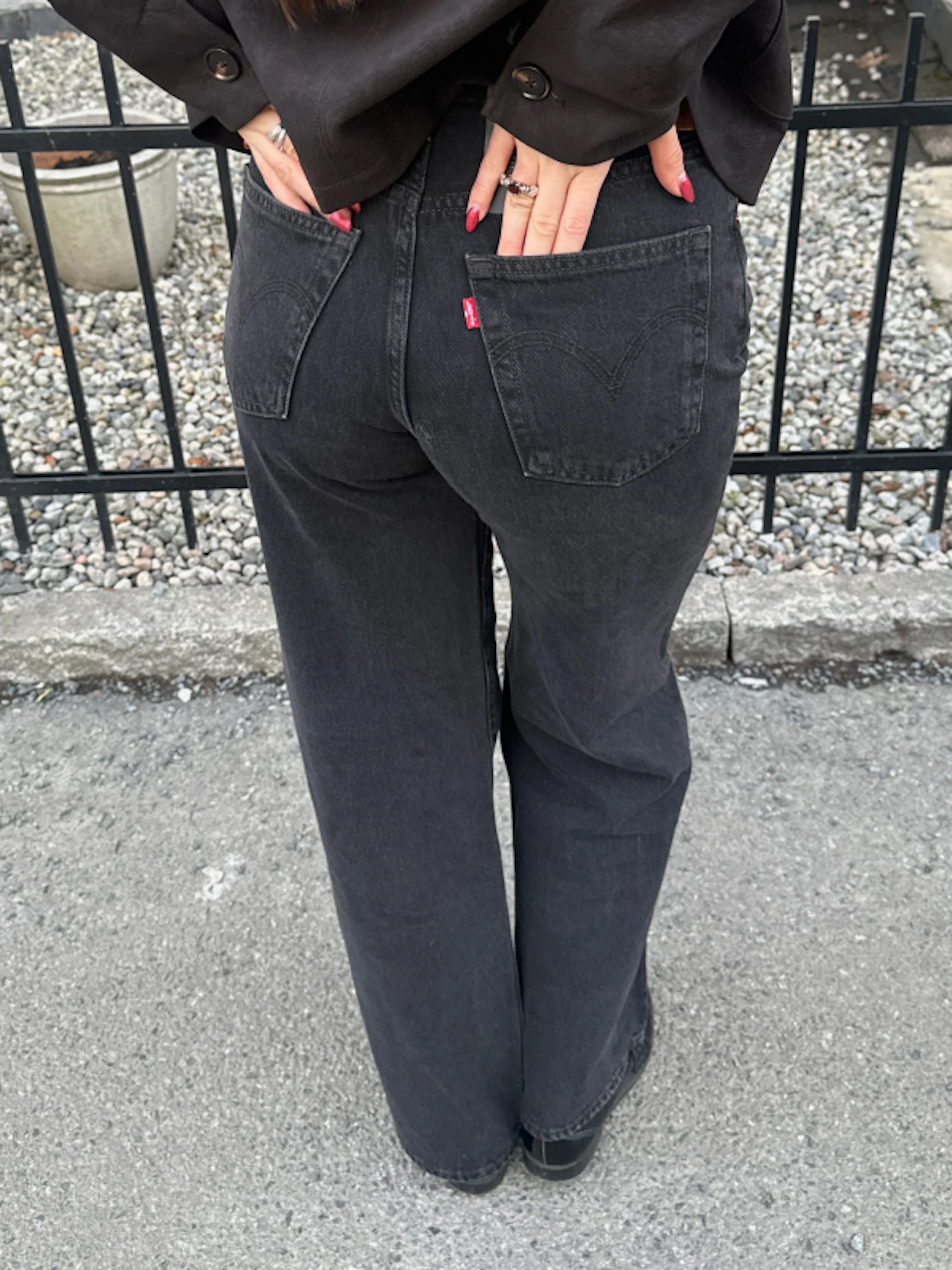 Person sett bakfra iført høytlivs svarte Levi's-jeans med hendene i baklommene, stående på asfalt foran et lavt metallgjerde og en grusfôret plantekum.