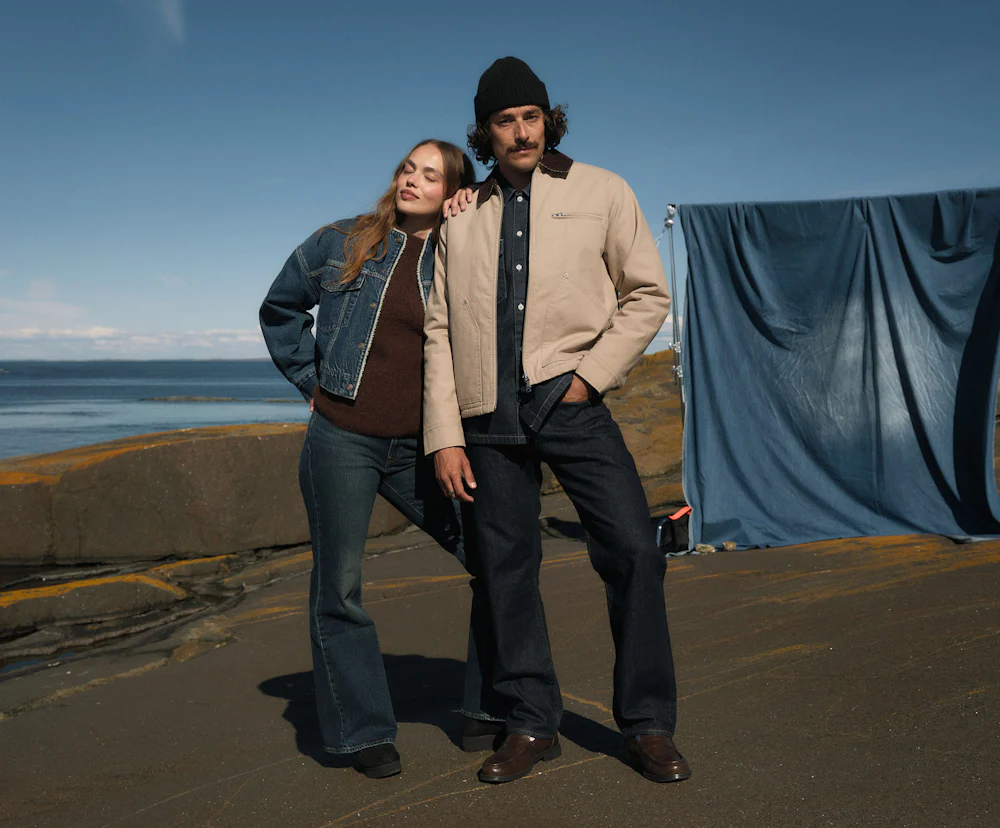To personer i casual denimantrekk poserer utendørs ved havet. Hun lener seg mot ham under blå himmel.