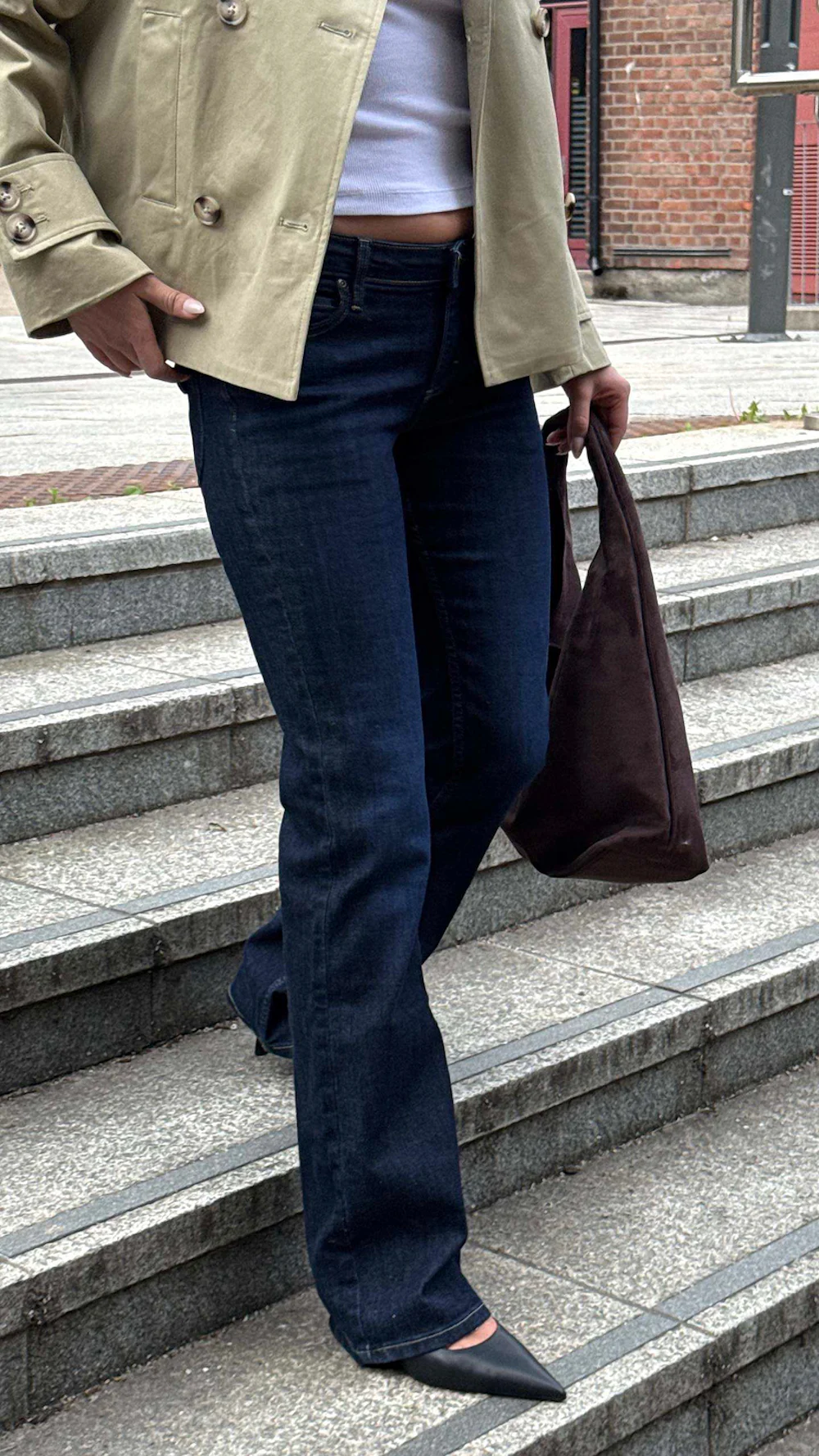 Person i mørkeblå jeans, beige jakke og svarte hæler, bærer en mørk brun veske på utendørs trapper.