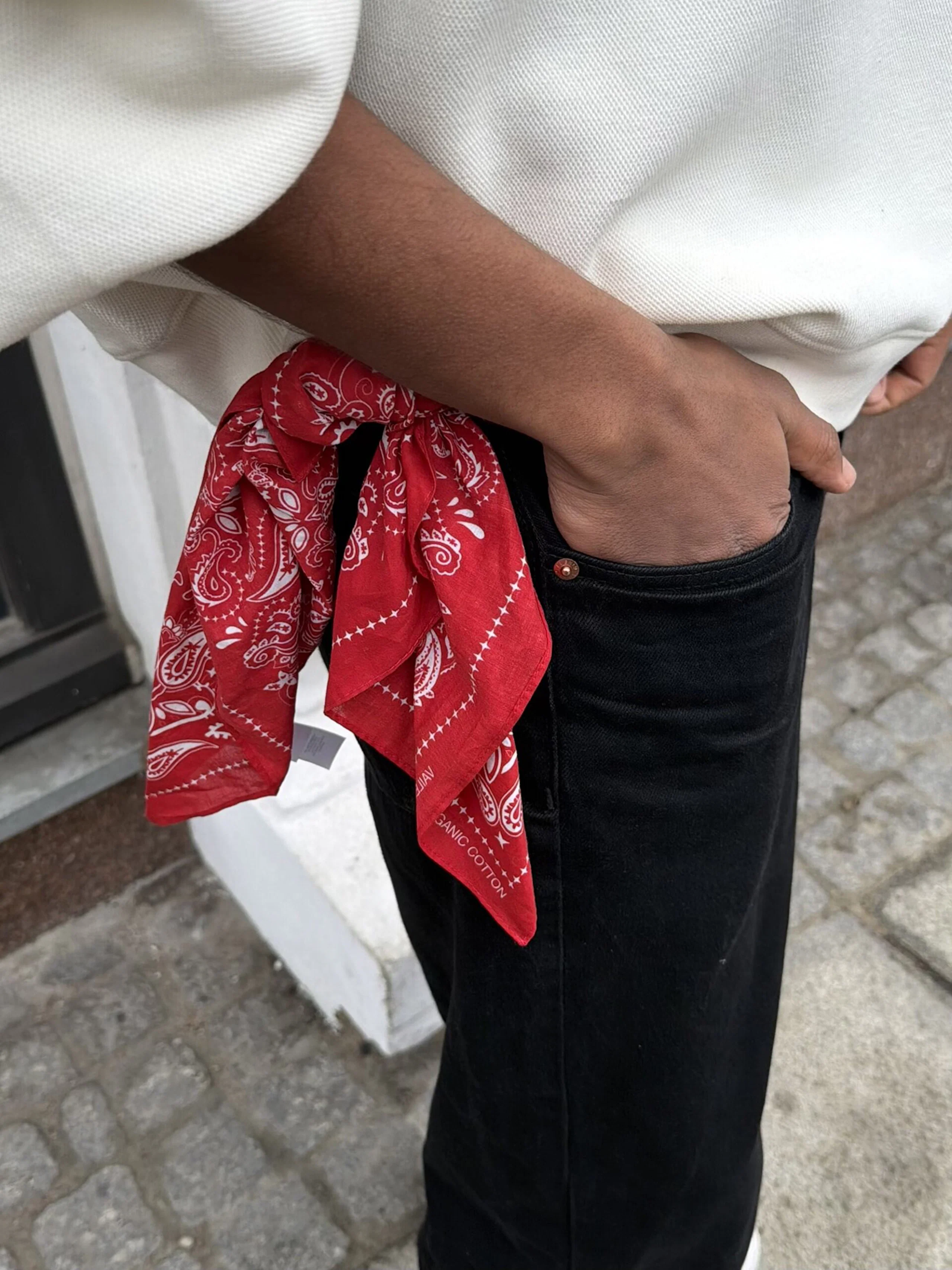 Röd bandana med vitt paisleymönster knuten i bältesbandet på svarta jeans, buren av en person i vit tröja.
