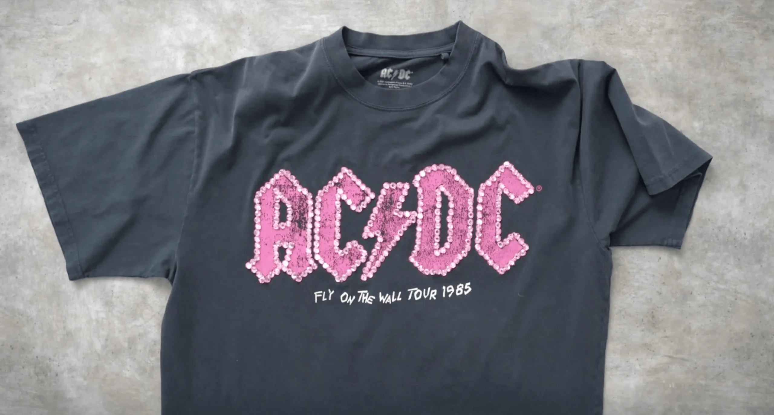 Svart AC/DC-turné-T-skjorte med rosa paljettert bandlogo og "Fly on the Wall Tour 1985"-tekst på en betongbakgrunn.