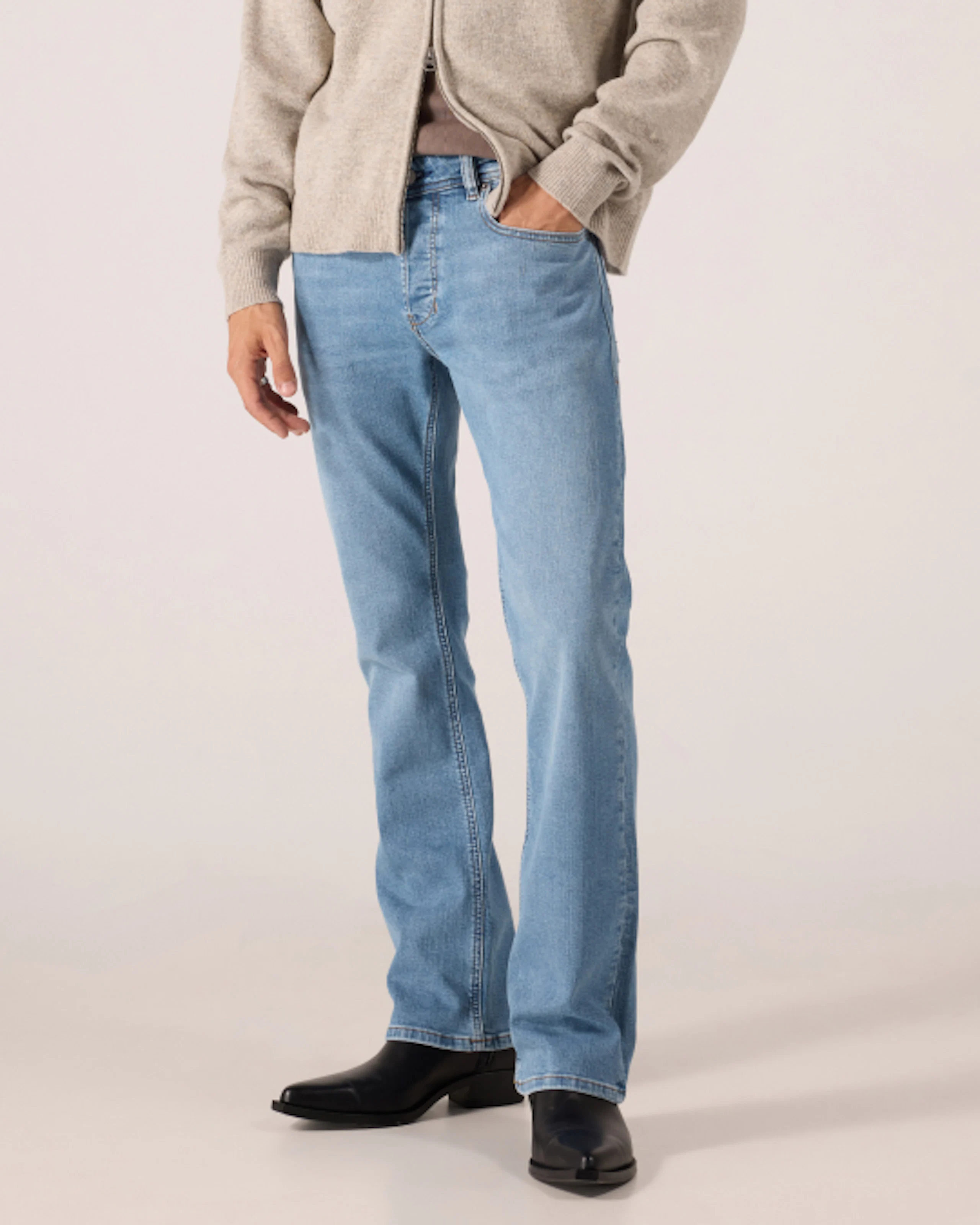 Mies, jolla on vaaleansininen kellohousut, beige vetoketjullinen pusero ja mustat cowboy-saappaat, vaaleanharmaan taustan edessä.