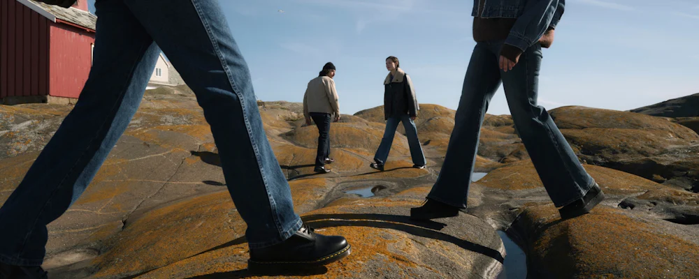 Fyra personer som går på orange lavbevuxna kustklippor; två i förgrunden visar närbild på jeansben och svarta kängor, två i bakgrunden bär jackor och jeans under en ljusblå himmel nära röda sjöbodar.