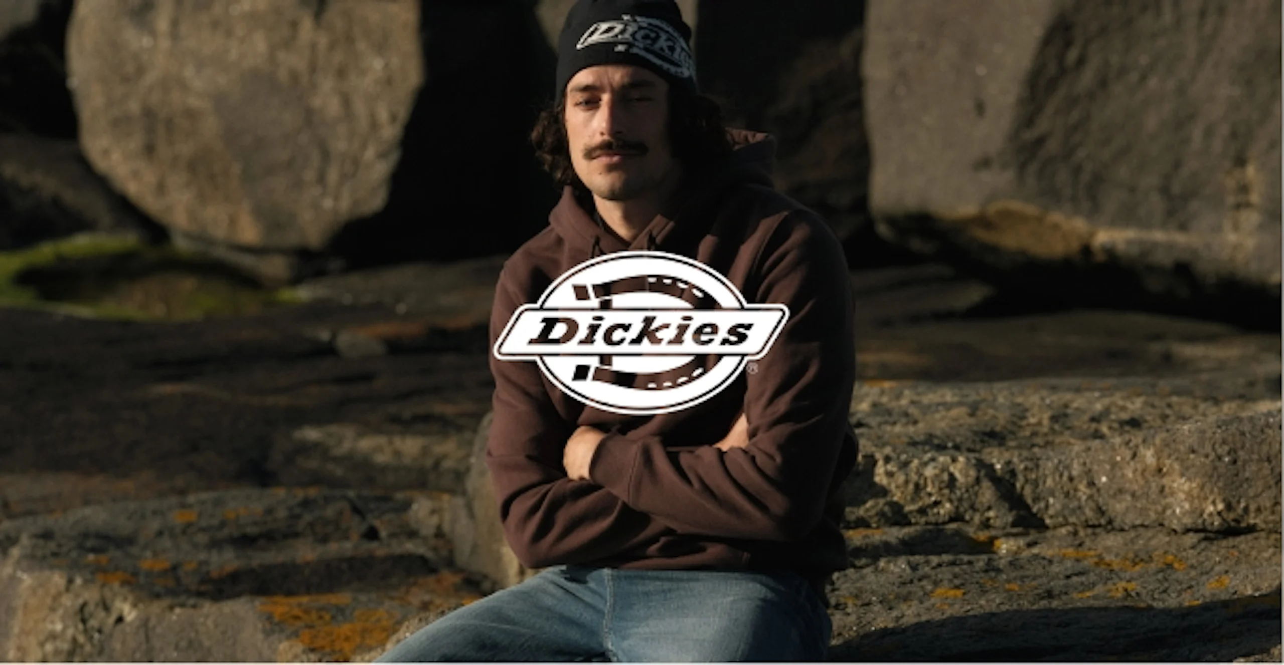 Nuori mies istuu kallioilla yllään ruskea Dickies-huppari ja farkut. Päässä Dickies-pipo, kädet ristissä.
