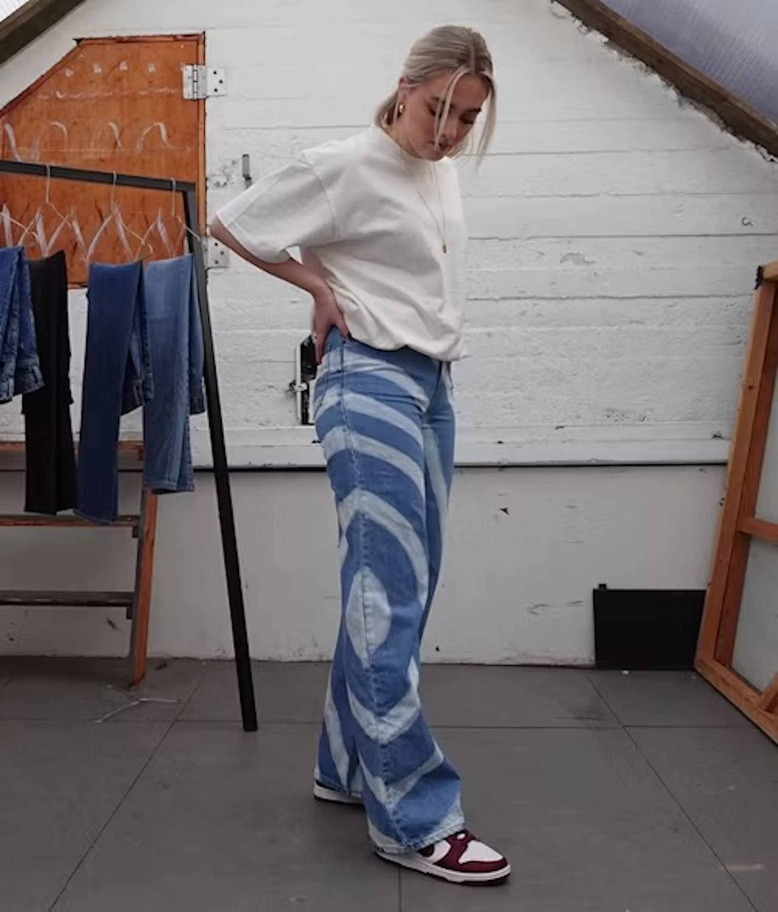 Ung kvinne står i studio iført hvit t-skjorte, blå tie-dye høytlivs vide jeans og røde-hvite joggesko, hendene på hoftene.