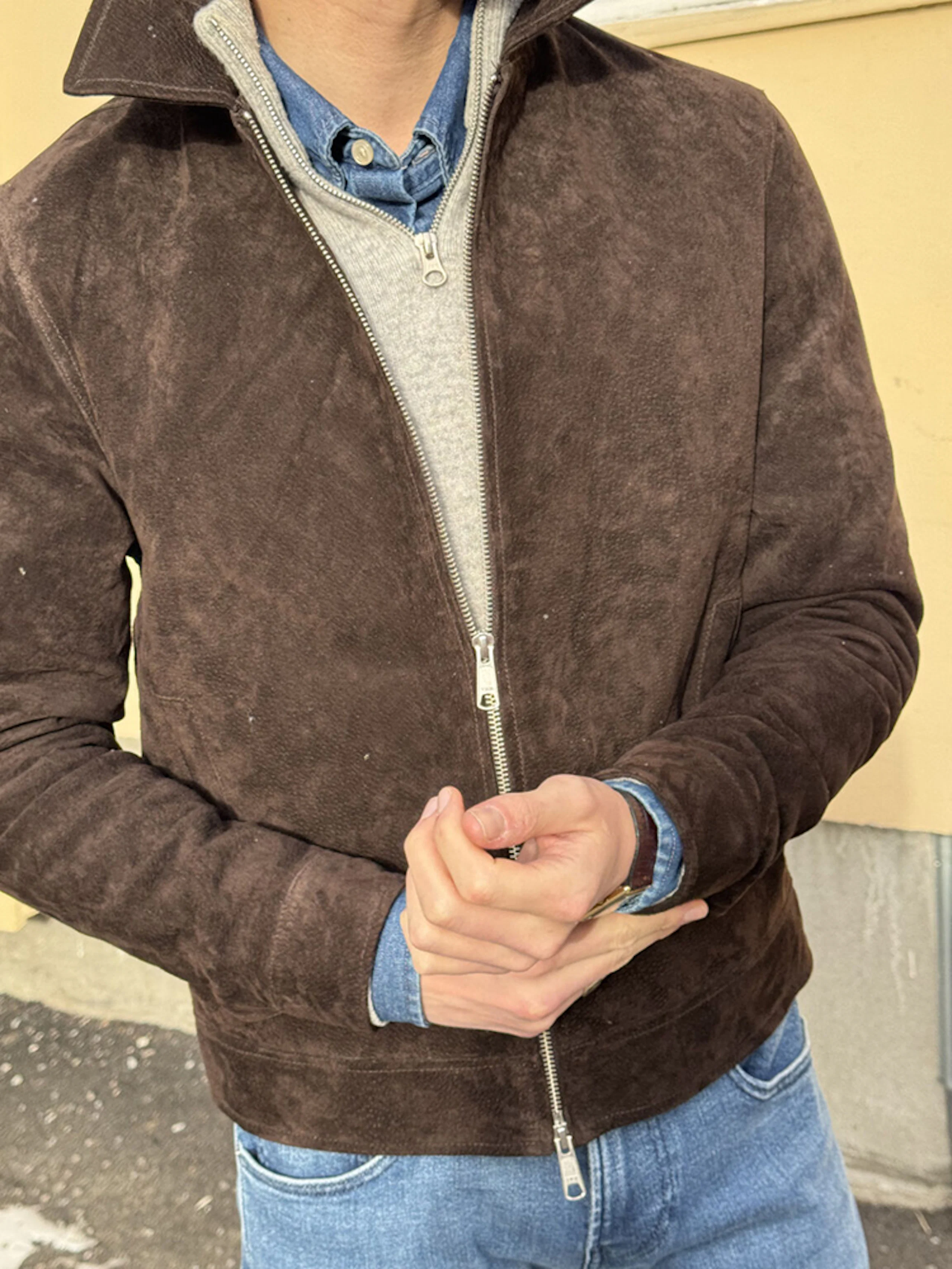 Man wearing a brown suede zip-up jacket over a gray quarter-zip sweater and blue denim shirt, hands clasped at waist and blue jeans visible.