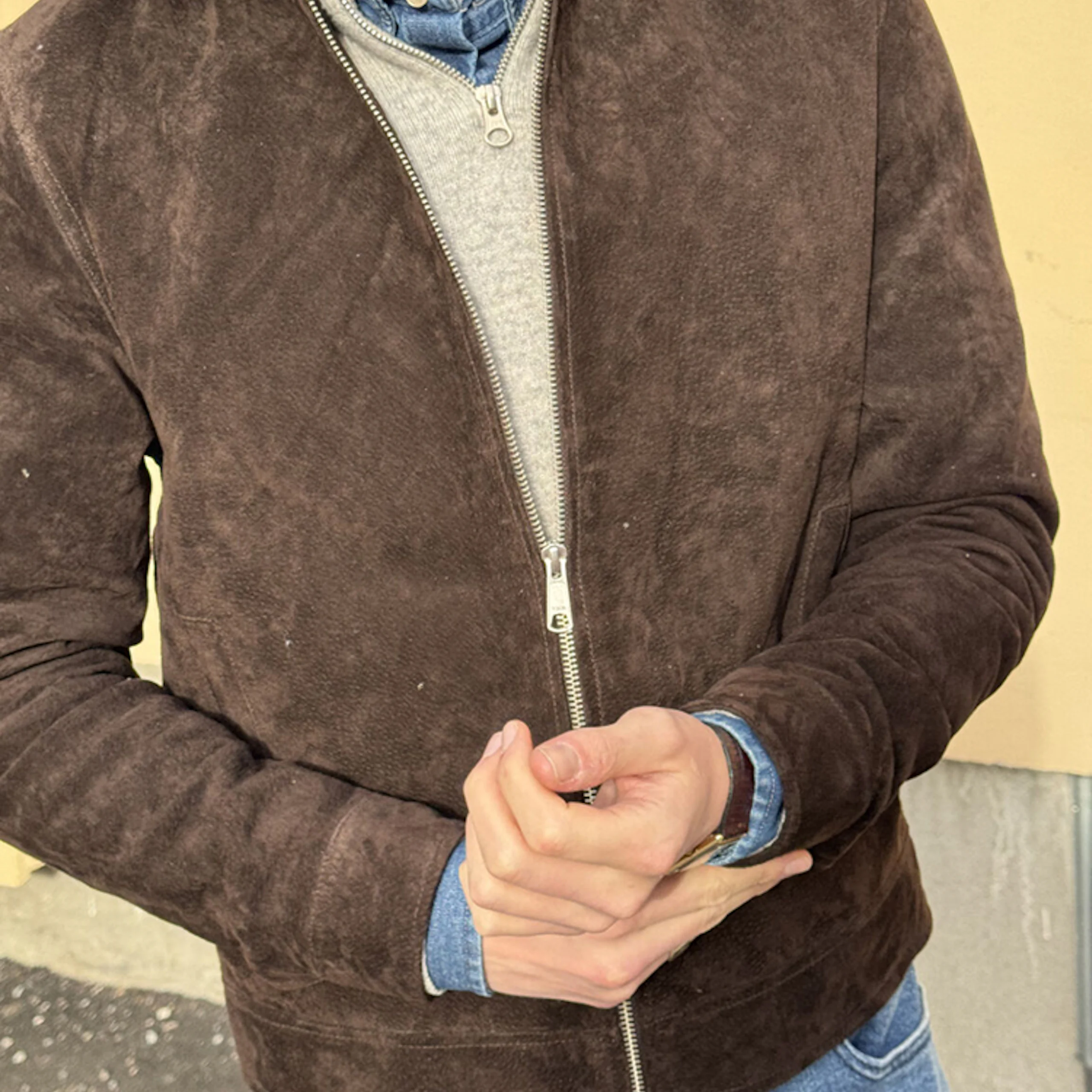 Man wearing a brown suede zip-up jacket over a gray quarter-zip sweater and blue denim shirt, hands clasped at waist and blue jeans visible.