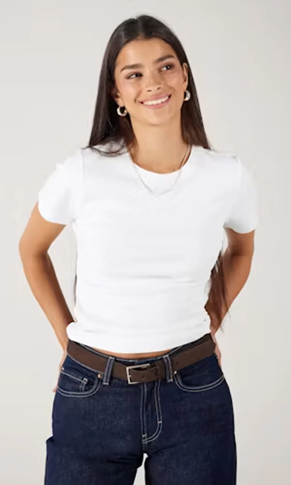 Ung kvinne iført en tettsittende hvit t-skjorte med korte ermer og mørkeblå jeans med brunt belte, smilende og ser til siden mot en ensfarget lys bakgrunn.