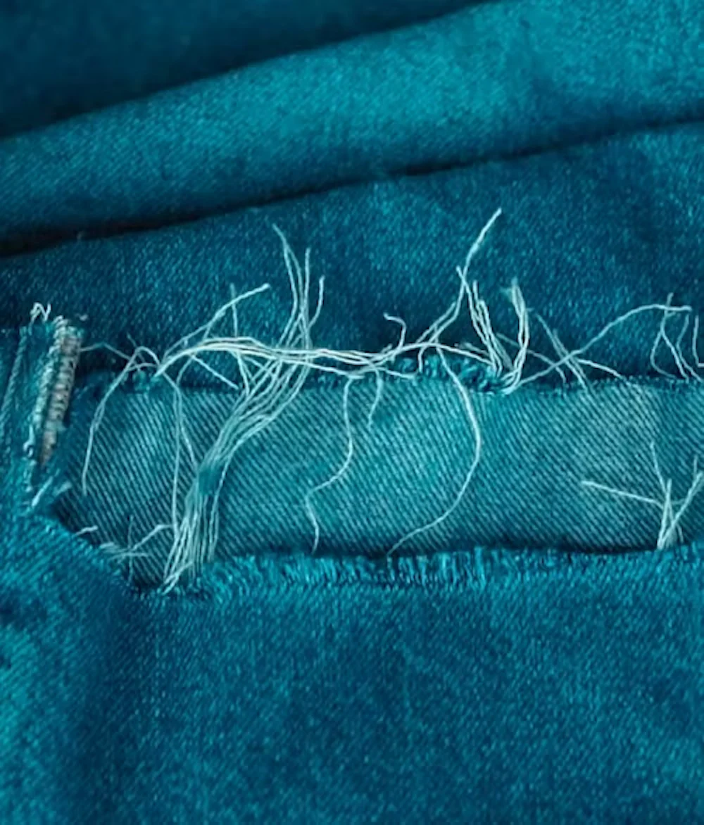 Nærbilde av slitt blågrønt denimlomme-søm med løse hvite tråder og synlig søm-detalj.