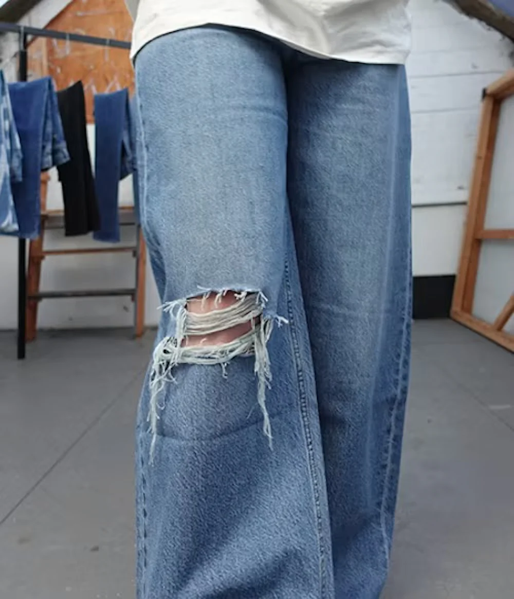 Nærbilde av lyseblå revne jeans med frynsete kneåpning, båret av en person som står i studio med hengende denim i bakgrunnen.