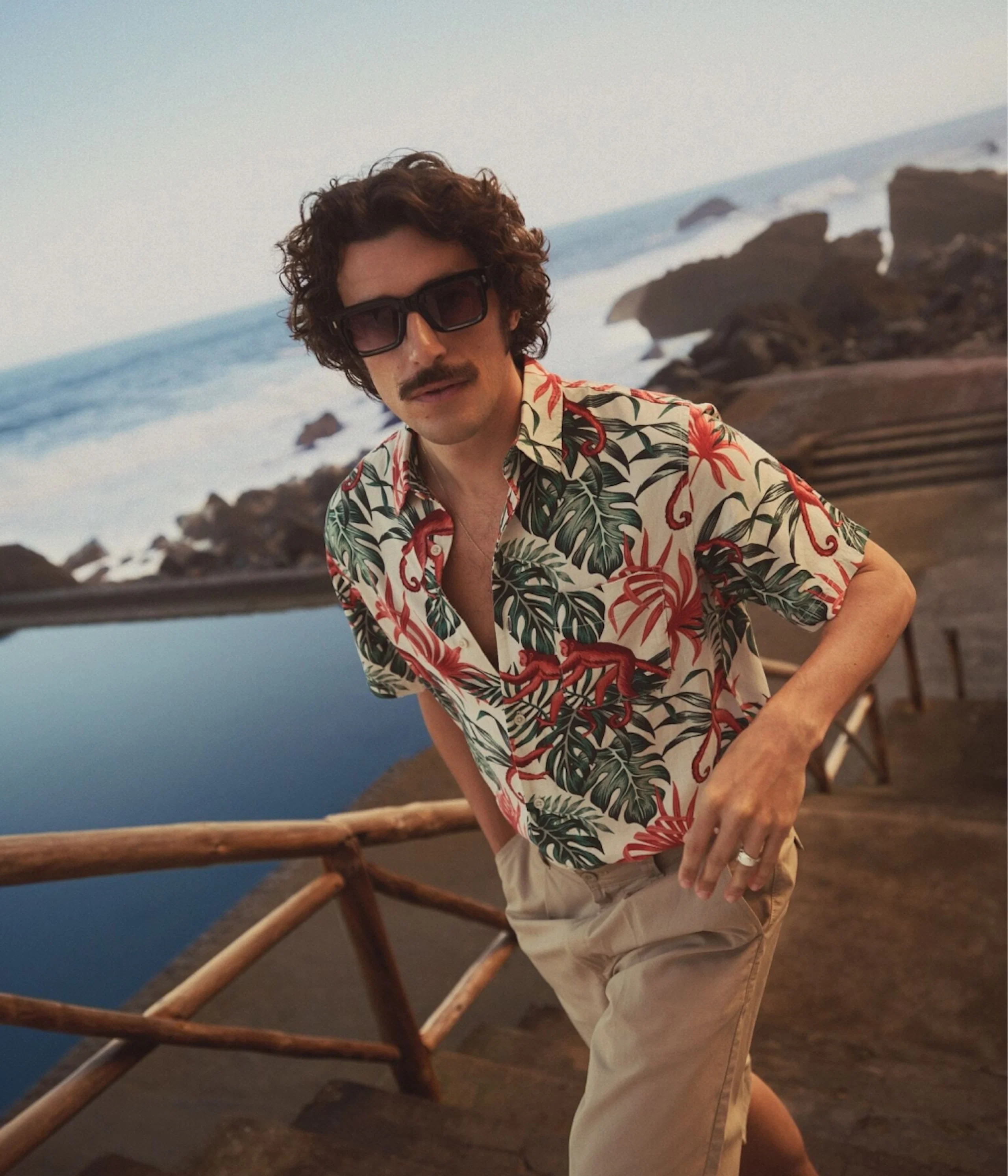 Mann med solbriller og tropisk skjorte med apemønster, beige shorts, lener seg mot et gjerde ved havet.