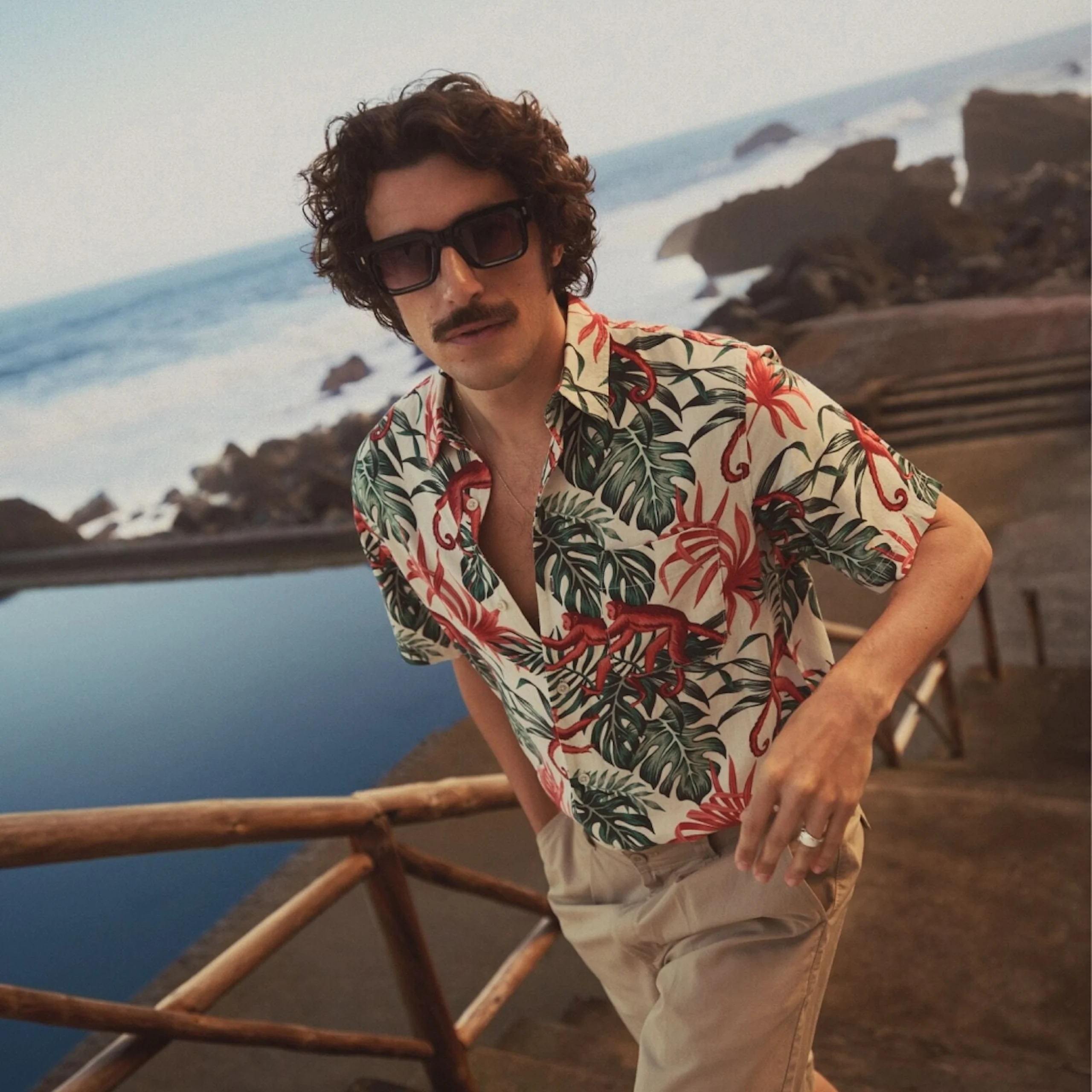 Mann med solbriller og tropisk skjorte med apemønster, beige shorts, lener seg mot et gjerde ved havet.