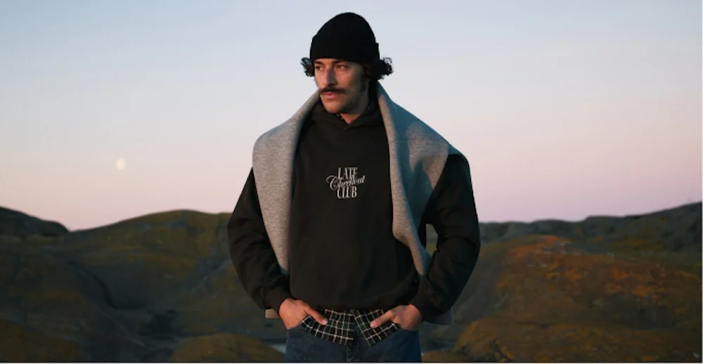 Man i svart hoodie med texten Late Checkout Club, grå tröja runt axlarna och svart mössa, utomhus vid skymning.
