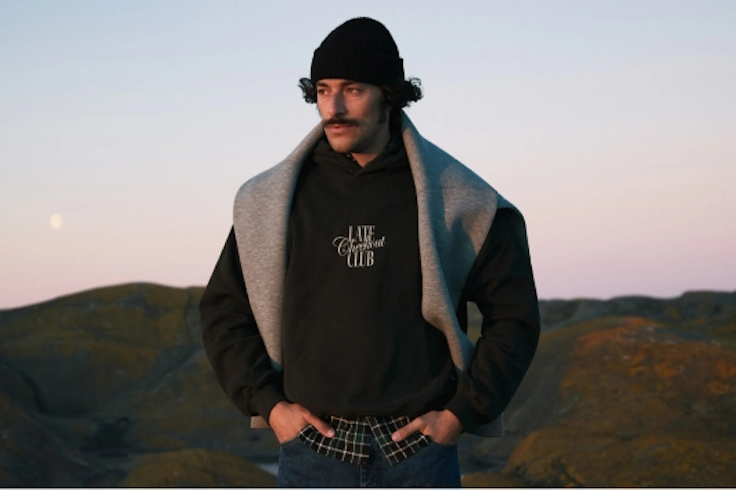 Man i svart hoodie med texten Late Checkout Club, grå tröja runt axlarna och svart mössa, utomhus vid skymning.