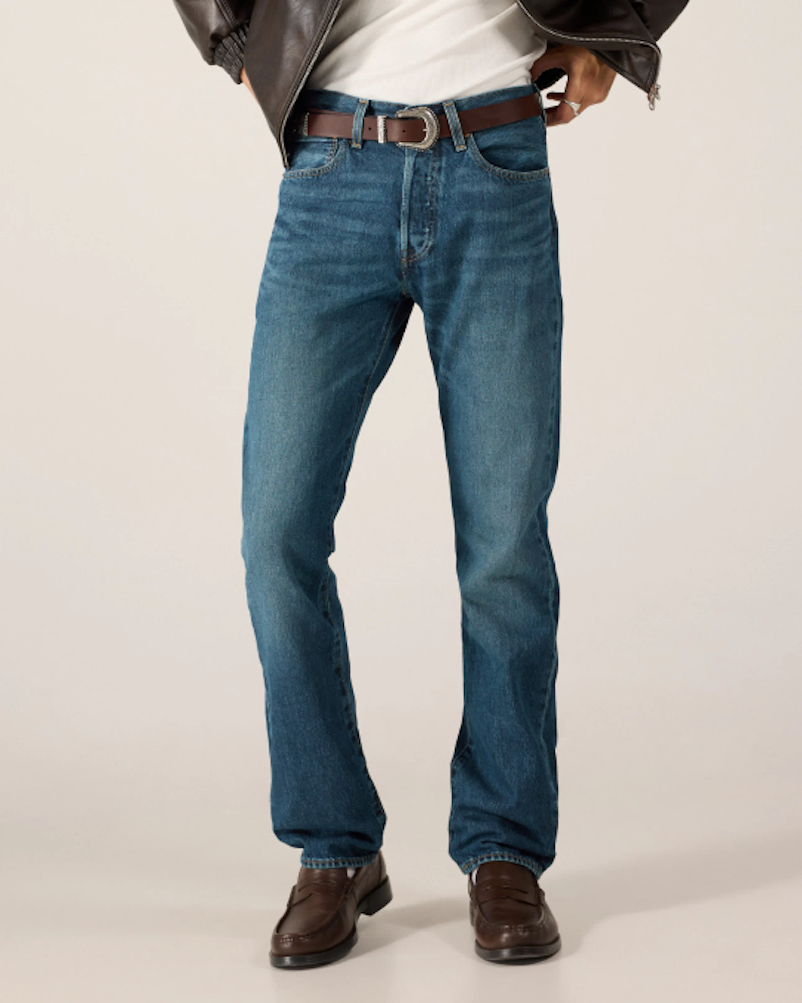 Mann iført mellomblå jeans med rett passform, brun belte og mørkebrune loafers, stylet med hvit t-skjorte og brun skinnjakke.