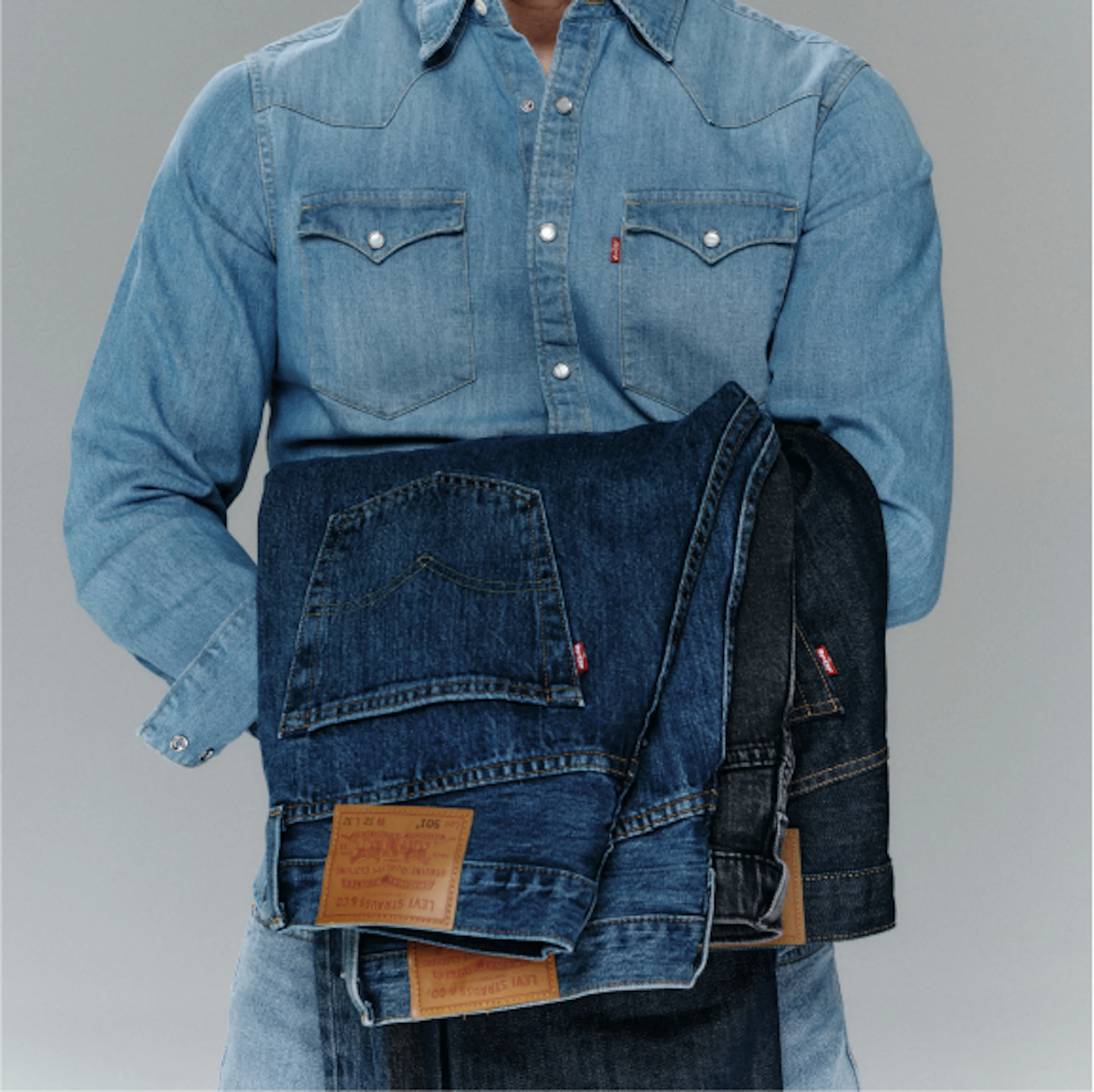 Henkilö sinisessä farkkupaidassa pitää sylissään useita taiteltuja Levi's-farkkuja eri sinisen sävyissä.