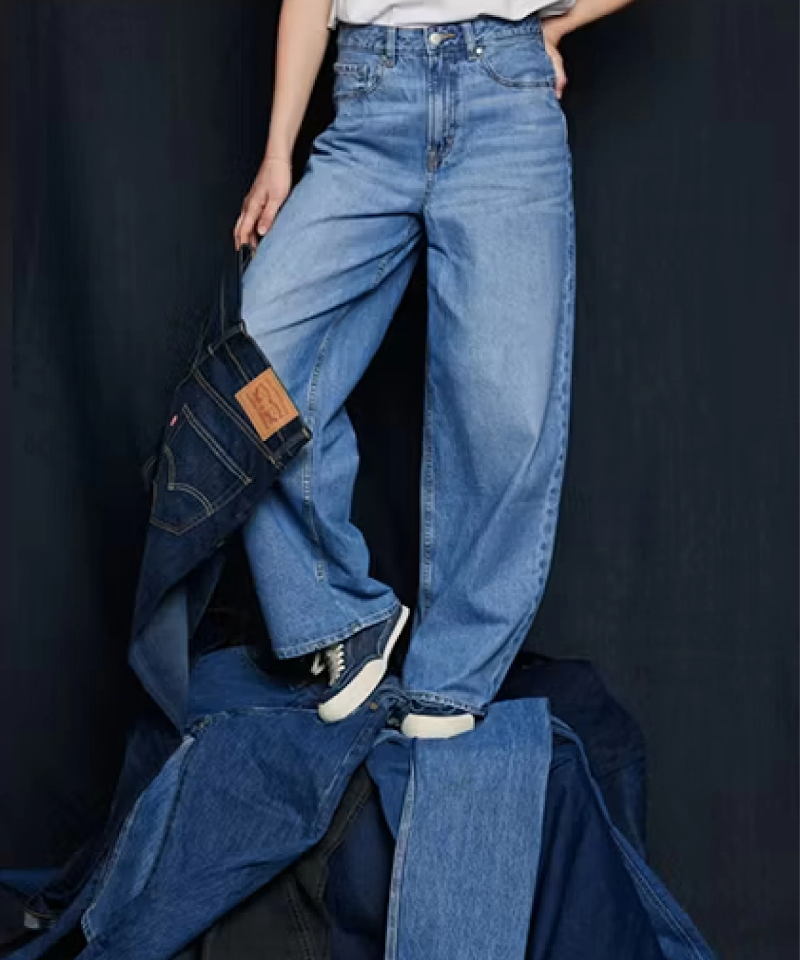 Nainen yllään vaaleansininen korkeavyötäröinen Levi's wide leg -farkku ja tennarit, ympärillä eri sinisiä farkkuja.