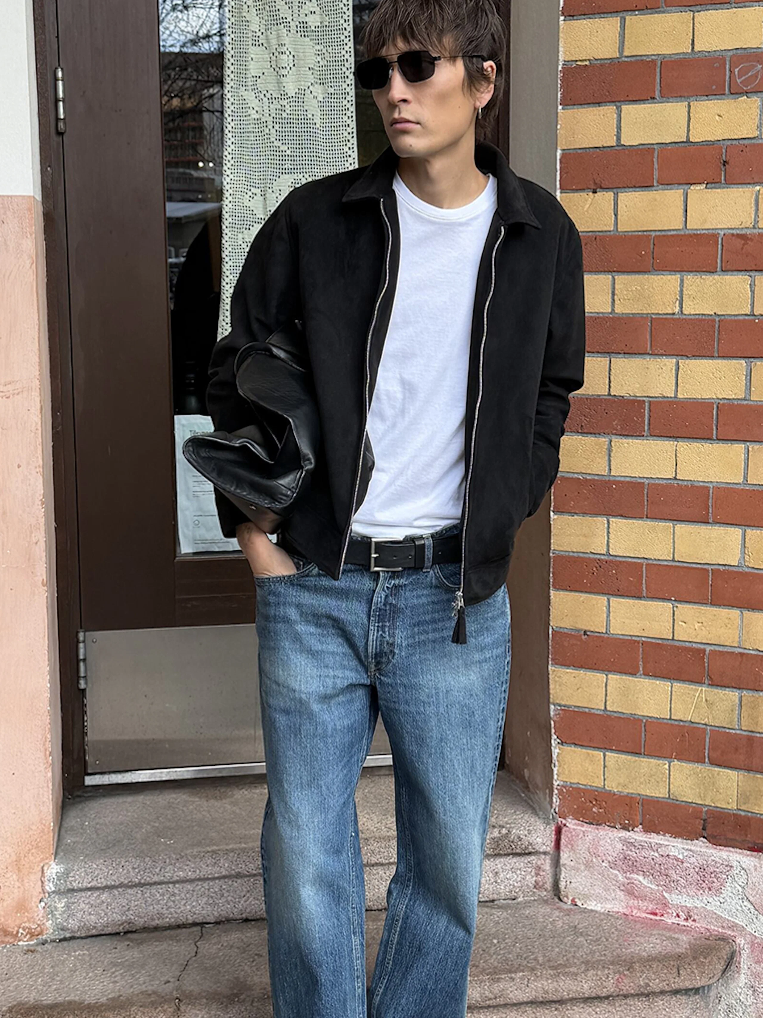 Man i svart jacka, vit t-shirt och blå jeans står framför en dörr vid en tegelvägg. Han bär solglasögon.