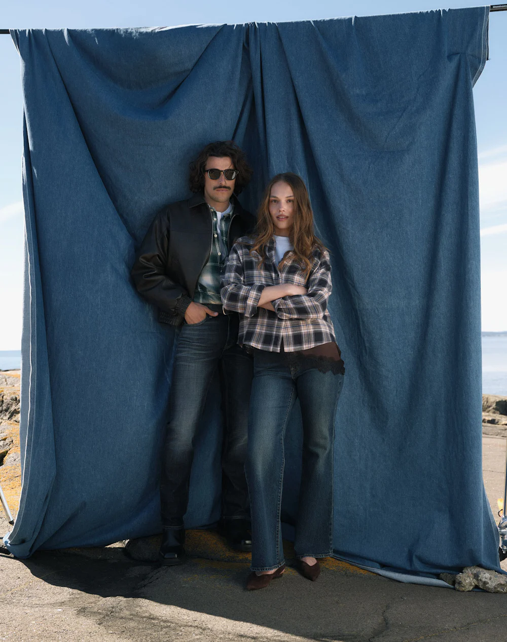 En man och kvinna i jeans och rutiga jackor poserar framför en blå tygbakgrund vid havet.