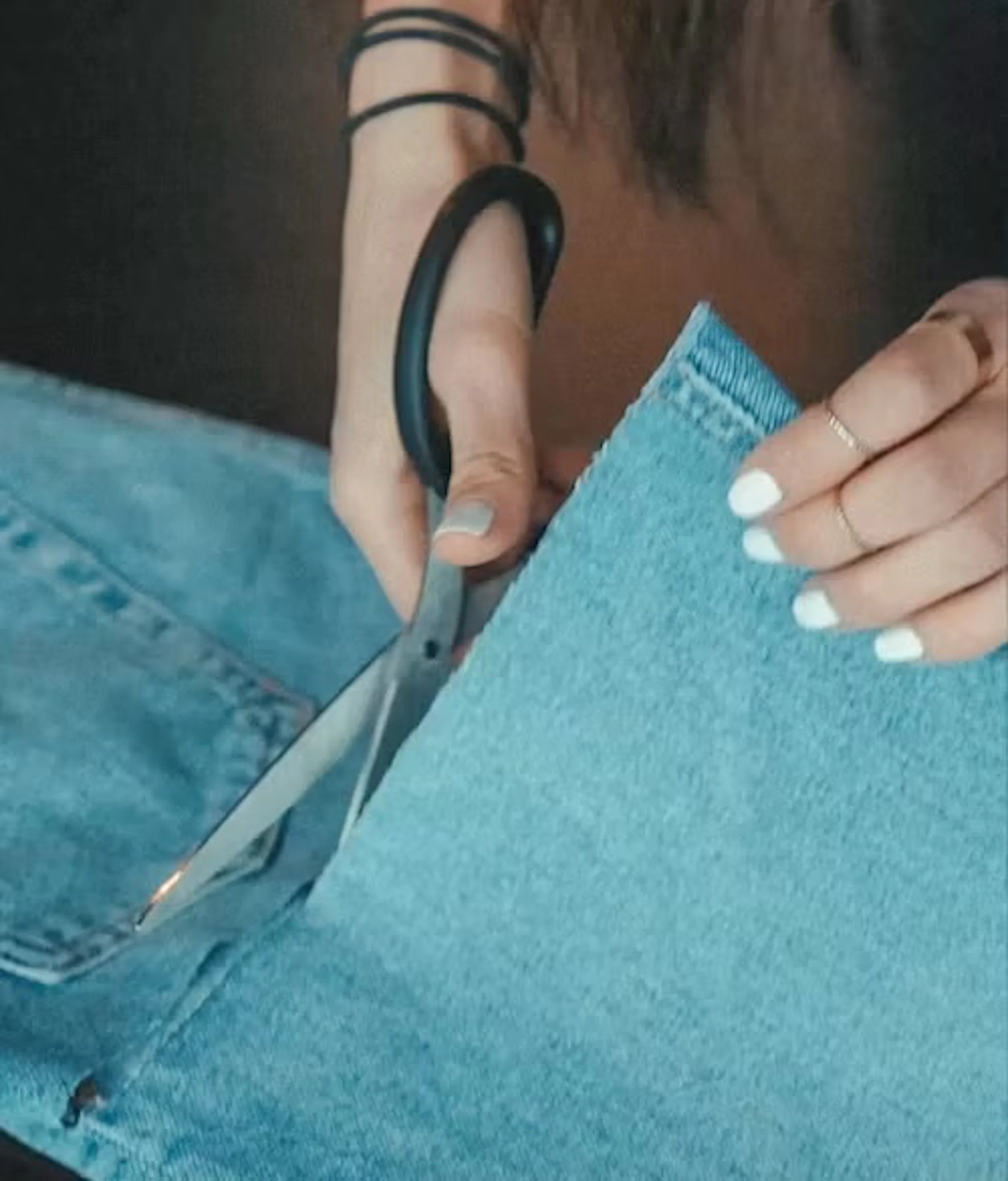 Hånd med hvitlakkerte negler som bruker saks med svart håndtak til å klippe lyseblå denimstoff, mens den andre hånden holder stoffkanten mot en mørk bakgrunn.