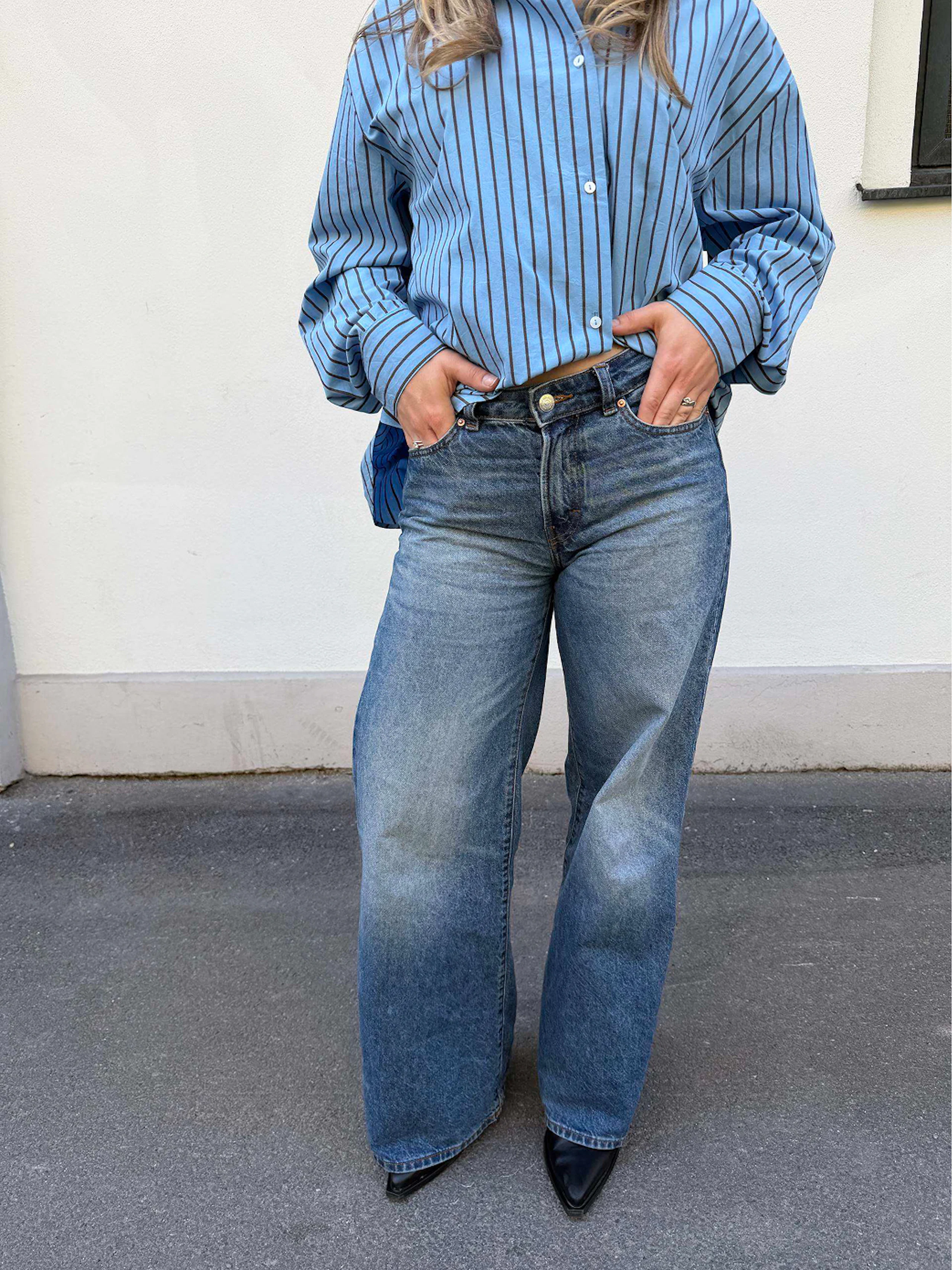 Kvinne iført blå stripete skjorte, vide blå jeans og svarte spisse sko, med hendene i lommene.