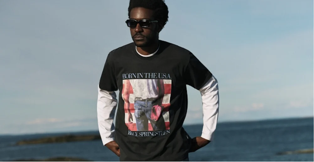 Man i svart Bruce Springsteen "Born in the USA" t-shirt och solglasögon står vid havet.