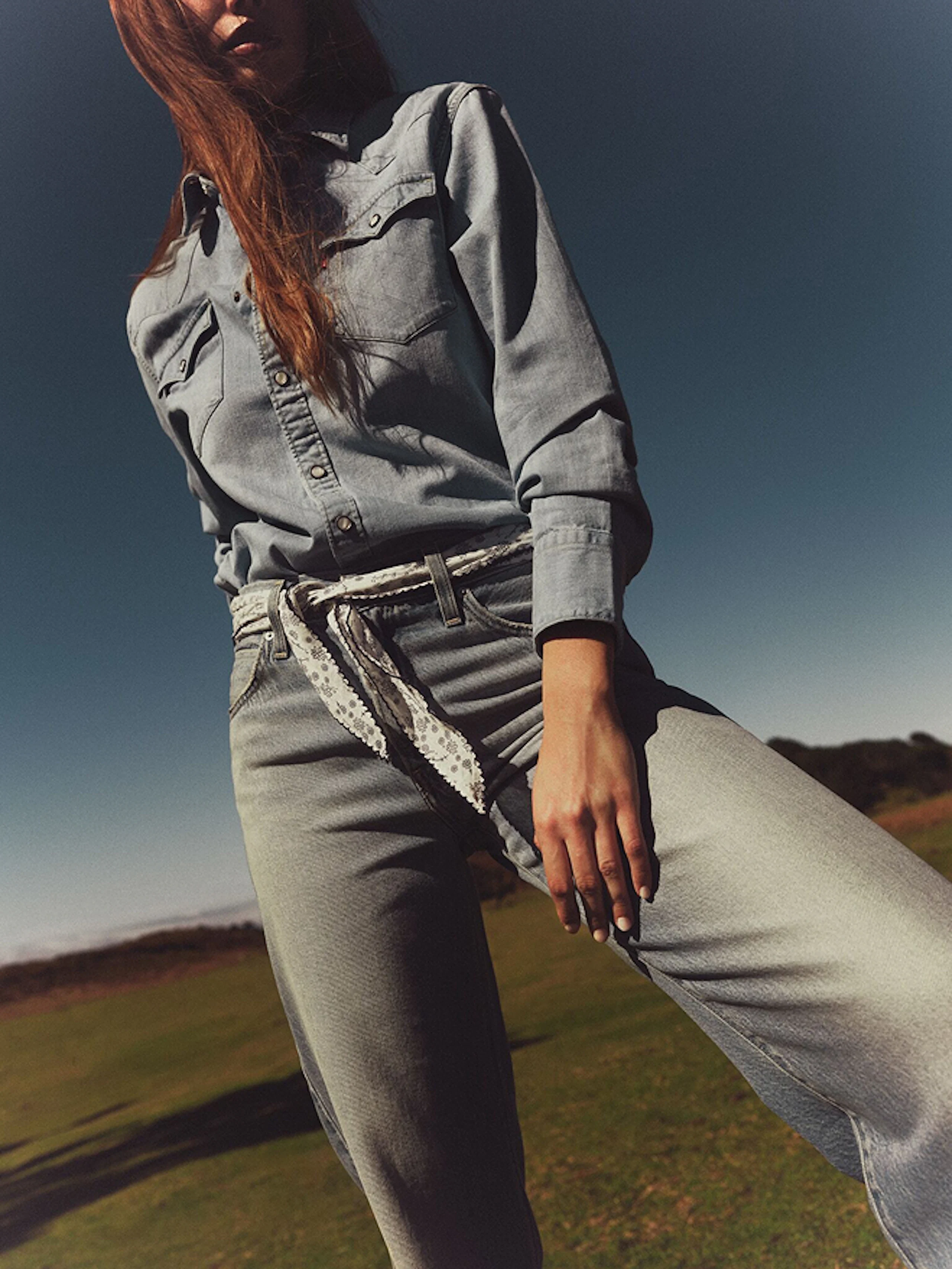 Woman wearing light denim shirt and jeans with a lace belt, standing outdoors on grass under a clear blue sky, hand resting on thigh.