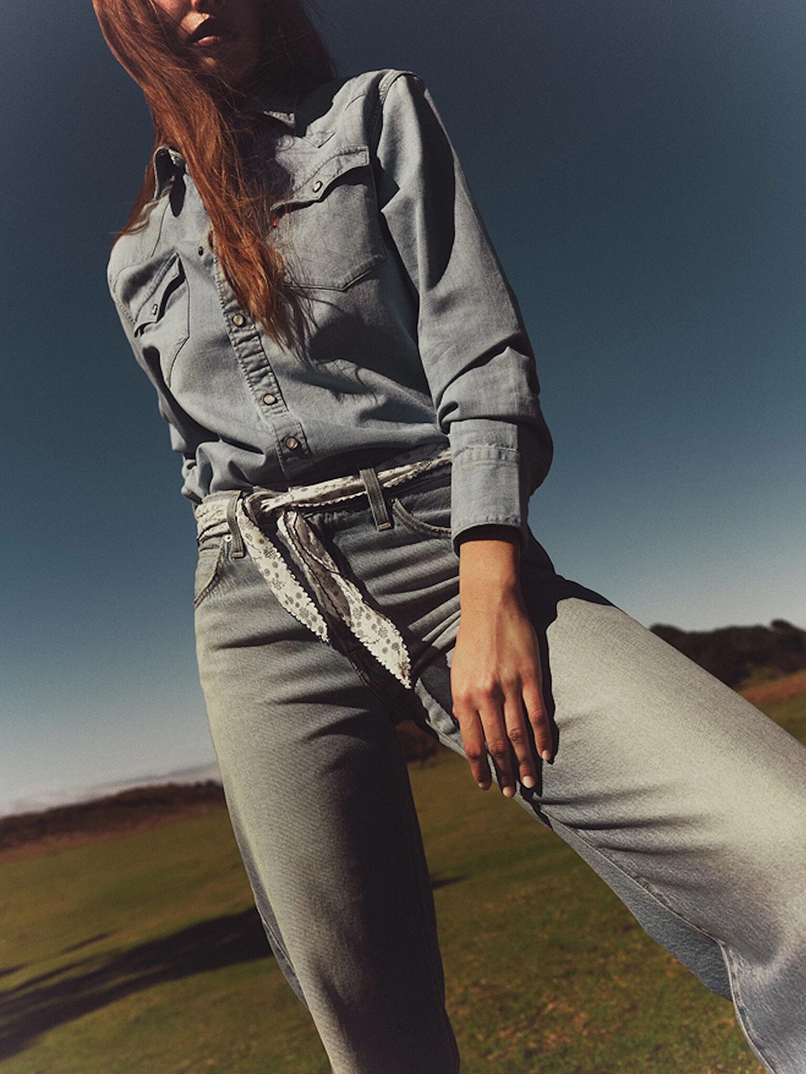 Woman wearing light denim shirt and jeans with a lace belt, standing outdoors on grass under a clear blue sky, hand resting on thigh.