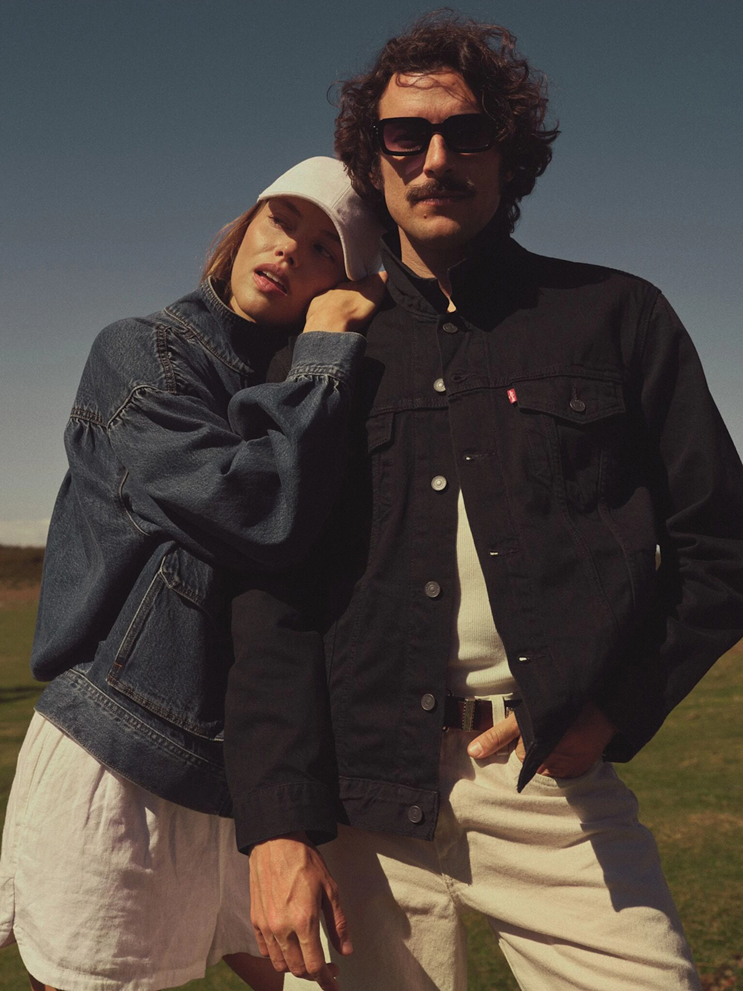 Young woman in white cap and denim jacket leans her head on a mustached man in sunglasses wearing a black denim jacket outdoors.