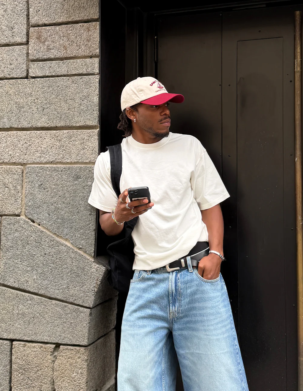 Ung mann i hvit T-skjorte, rødkantet baseballcap og vide jeans som lener seg mot en døråpning mens han holder en smarttelefon.