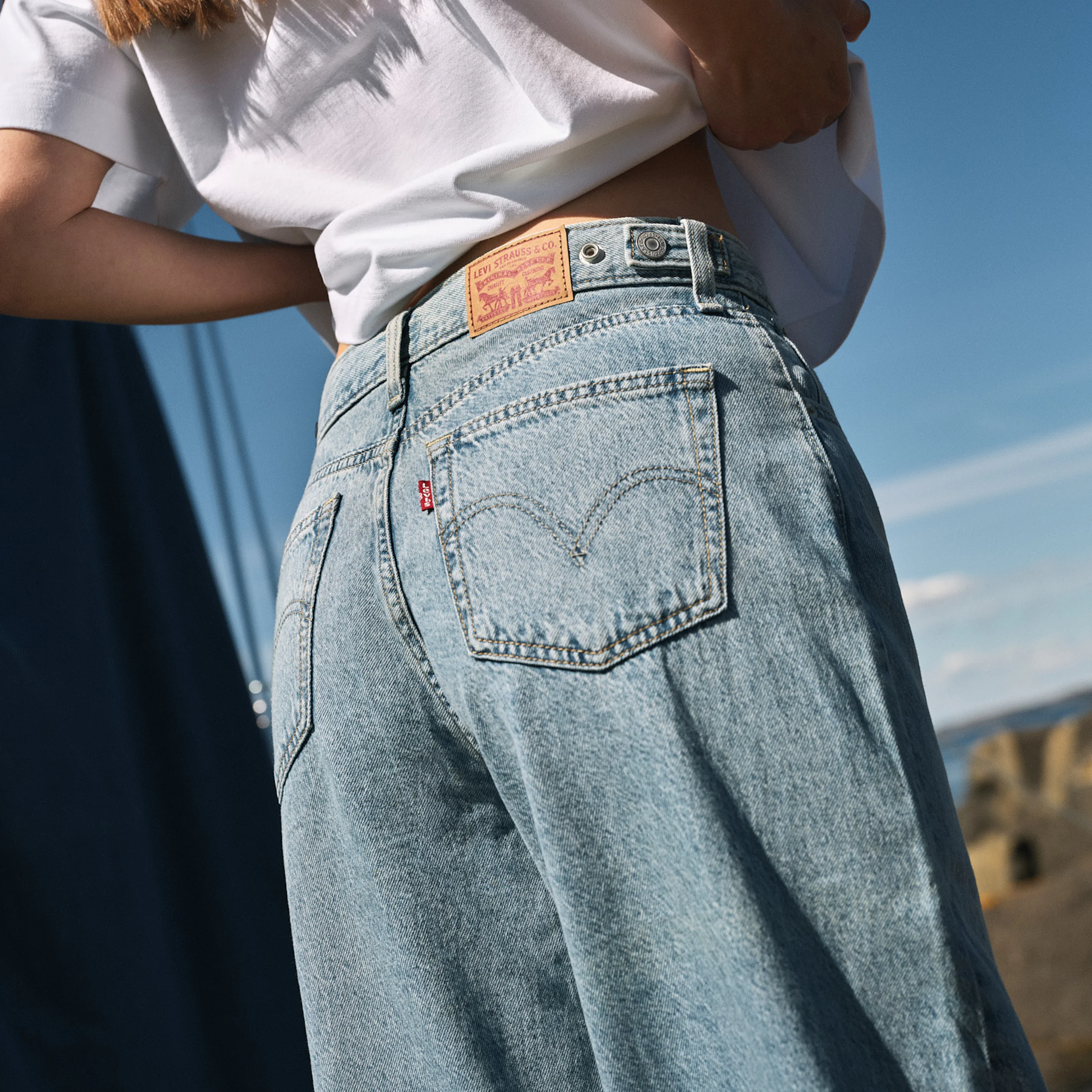 Nærbilde av en kvinne i lyseblå Levi's jeans med hvit t-skjorte, utendørs mot blå himmel.