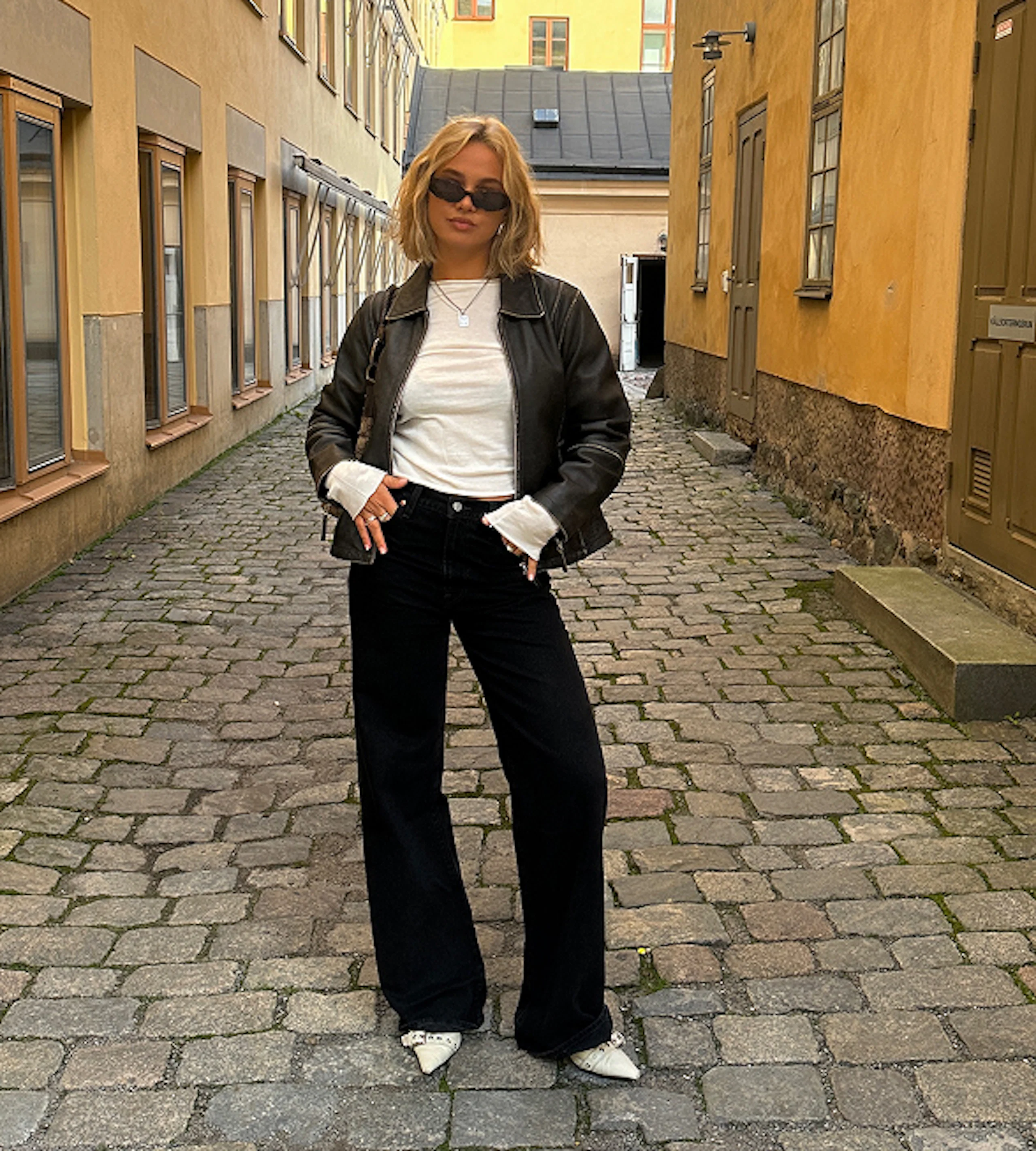 Ung kvinna i svart skinnjacka, vita t-shirt och svarta vida jeans står på kullerstensgata omgiven av gula byggnader.