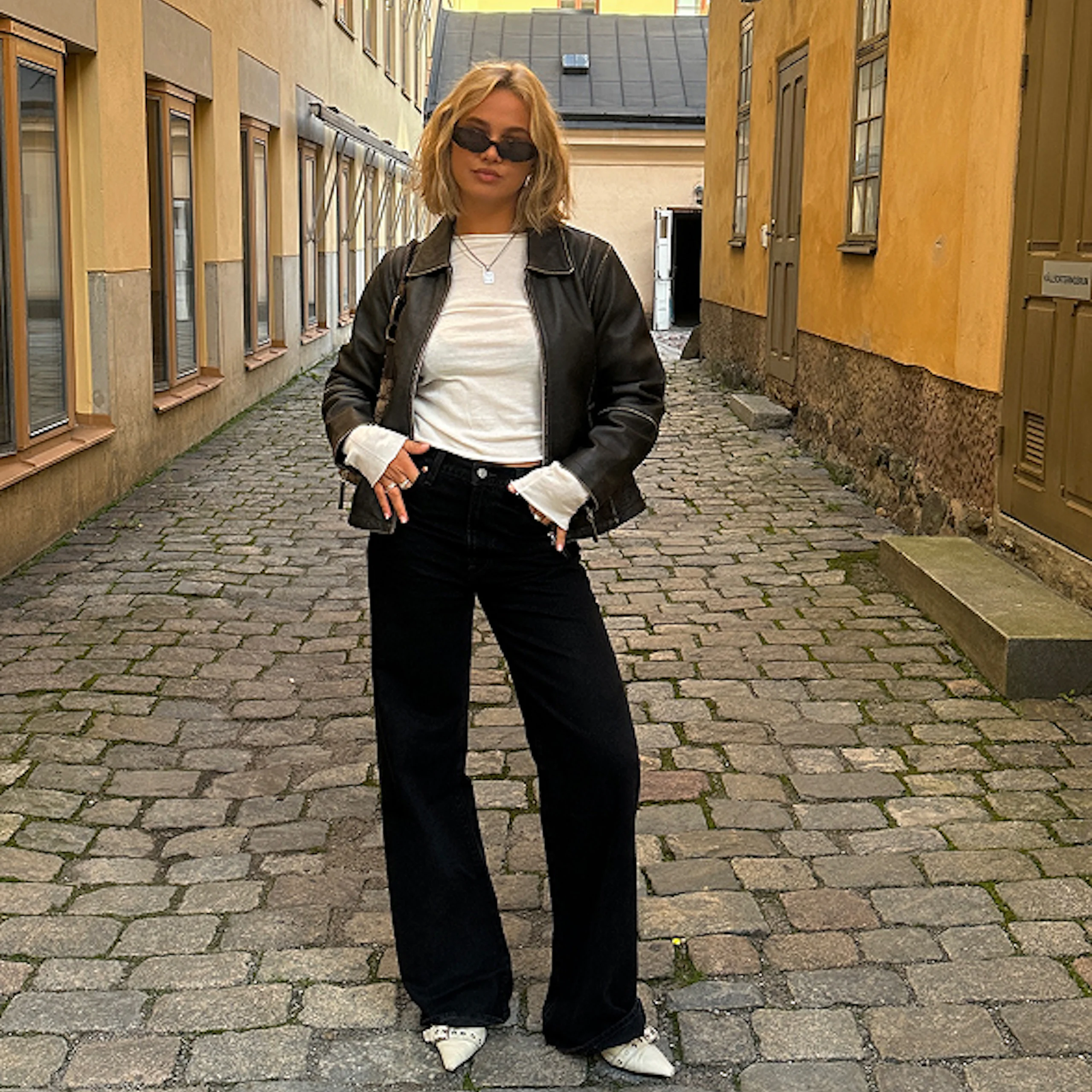 Ung kvinna i svart skinnjacka, vita t-shirt och svarta vida jeans står på kullerstensgata omgiven av gula byggnader.