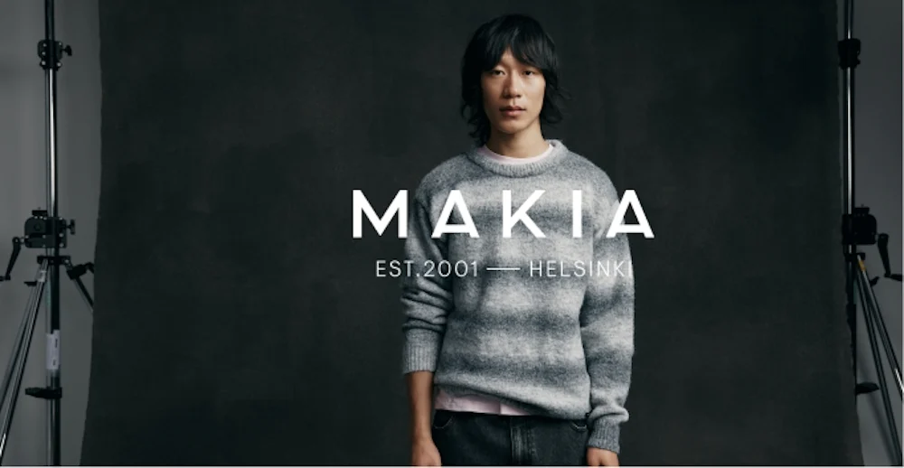 Makia-brändin mainos, jossa mies yllään harmaa raidallinen neule tummaa taustaa vasten. Teksti: Makia Est. 2001 Helsinki.