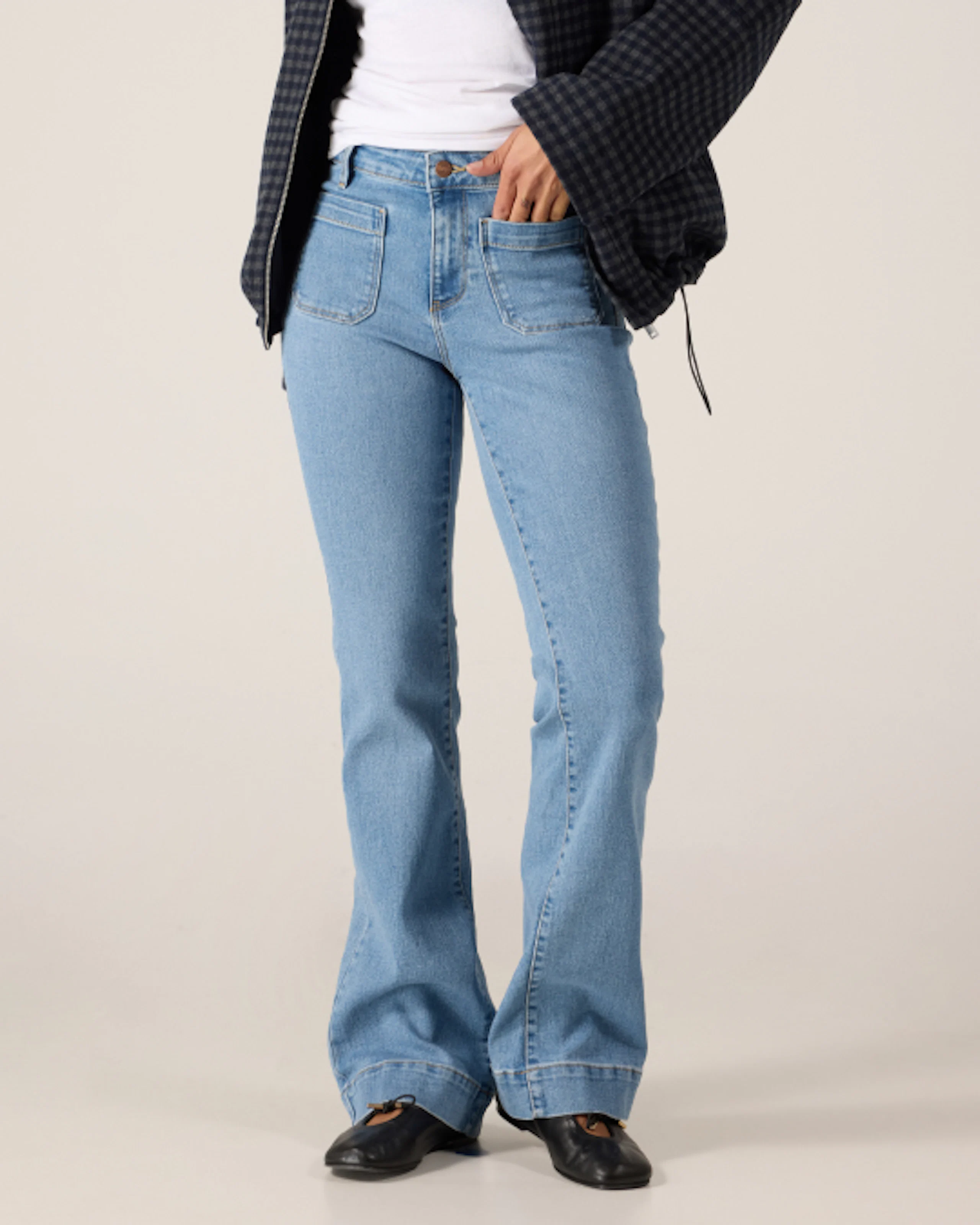Person i lyseblå utsvingede jeans, hvit topp og mørk rutete jakke, med svarte sko på nøytral bakgrunn.