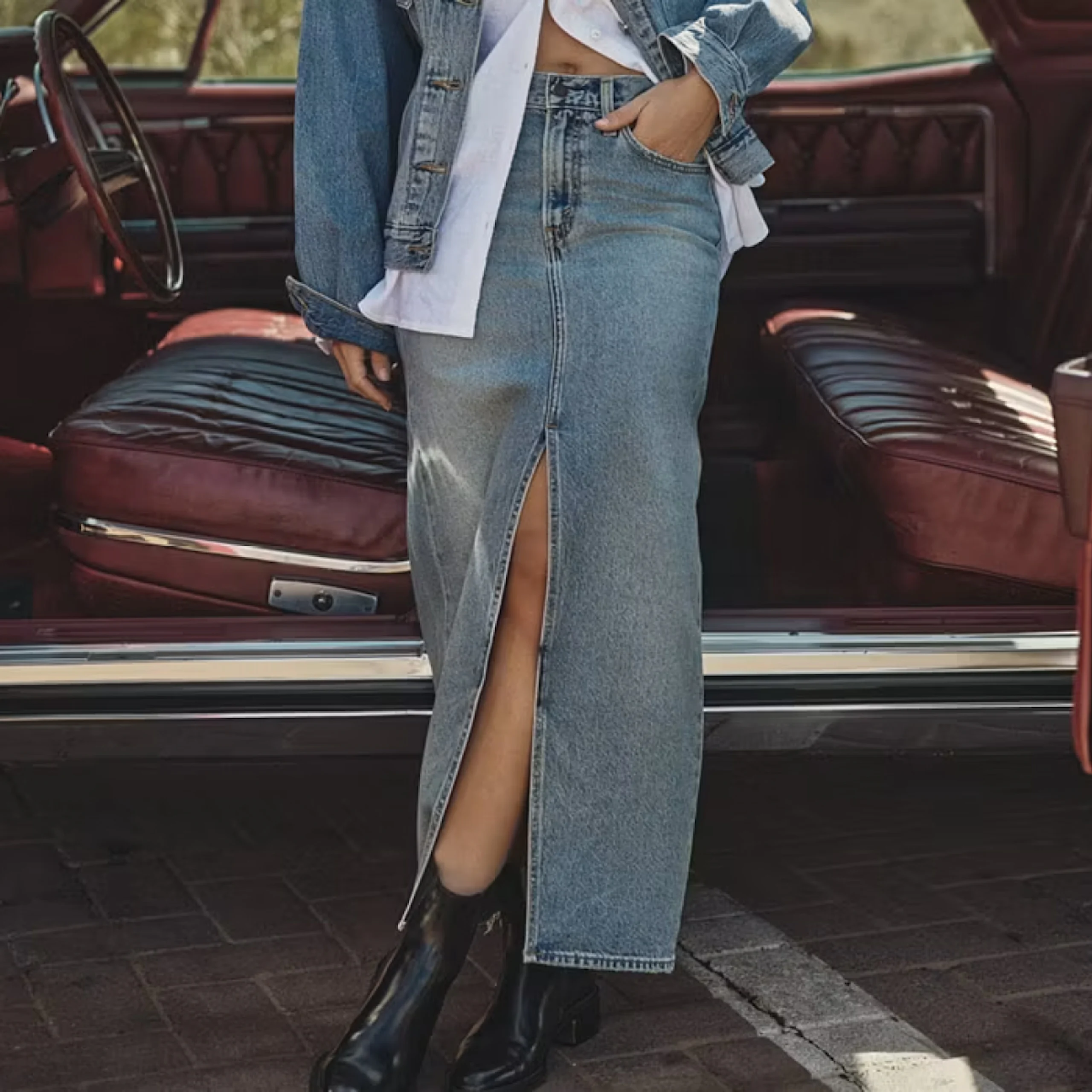 Kvinne i lang denimskjørt med splitt, denimjakke og svarte støvletter foran en klassisk bil med rødt skinninteriør.